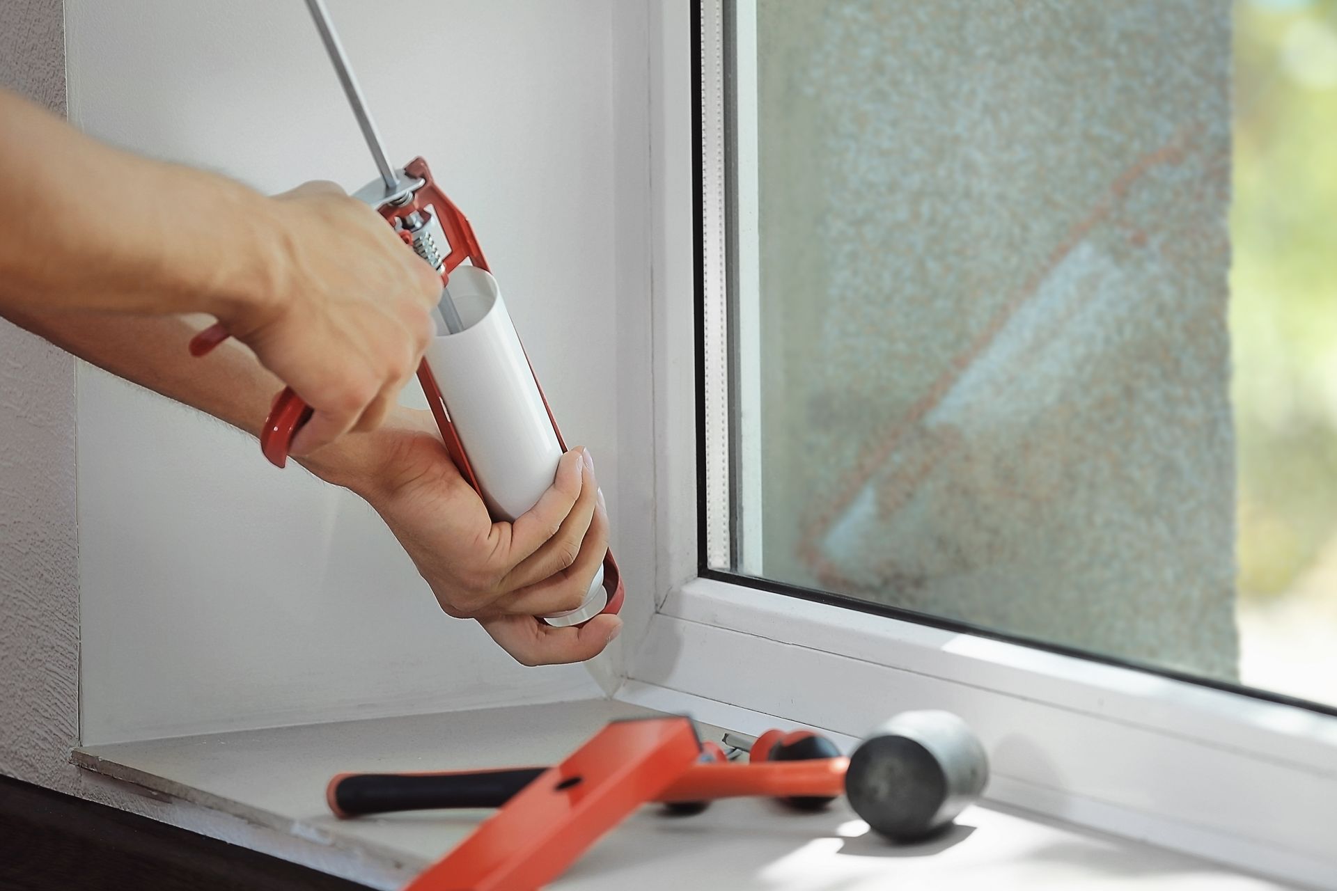 Hands applying caulk with a red caulking gun by a window sill