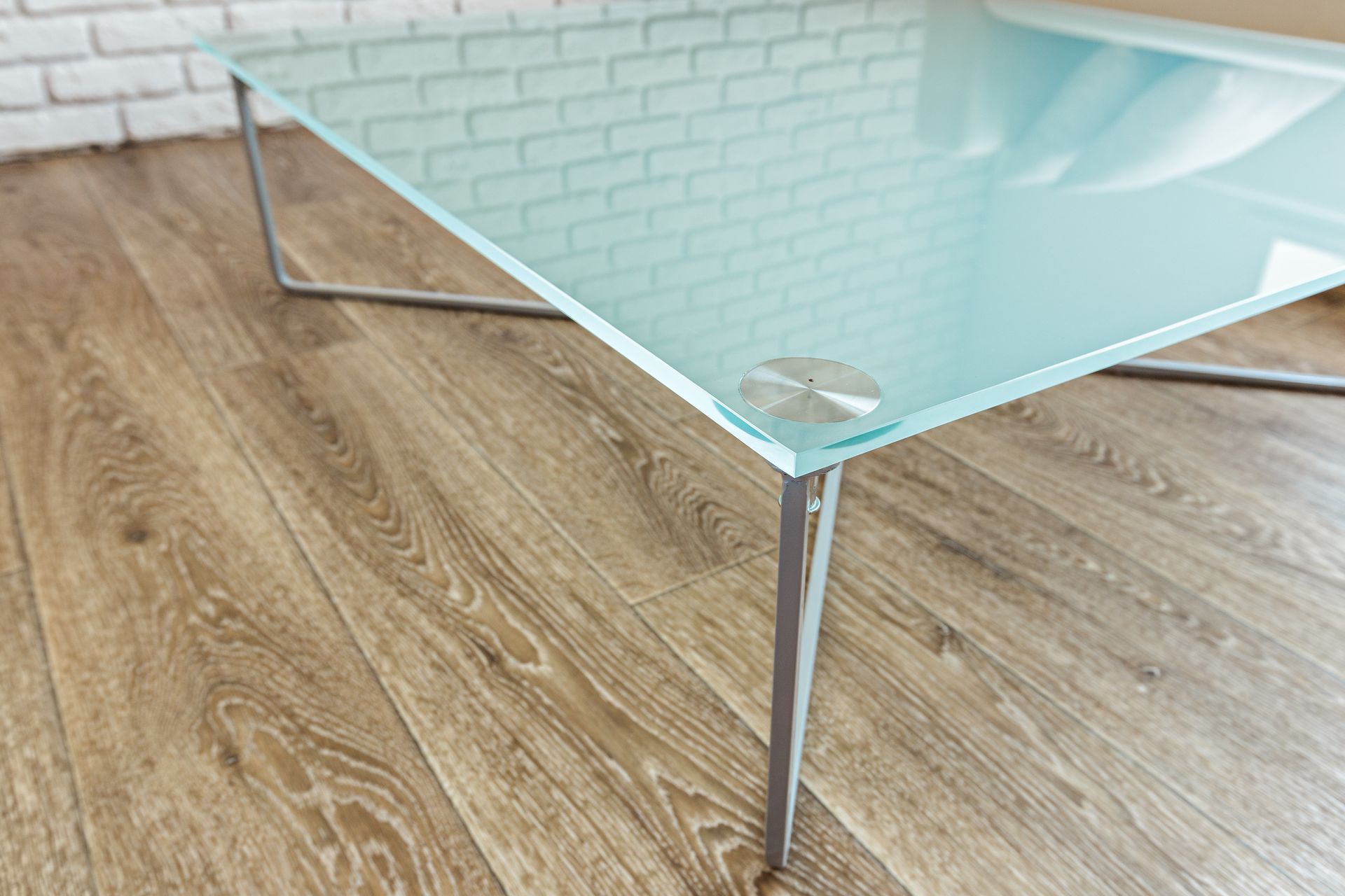 Glass coffee table with chrome legs on a light wood floor against a white brick wall