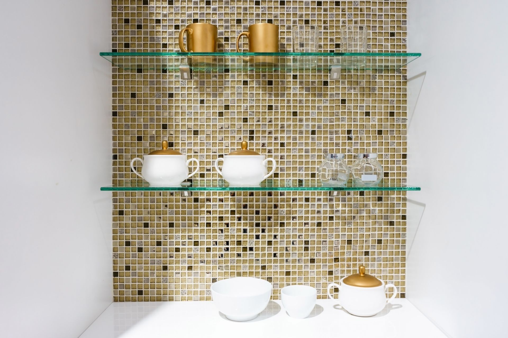 Minimalist display shelf with gold mosaic backing and white teacups on shelves and counter.