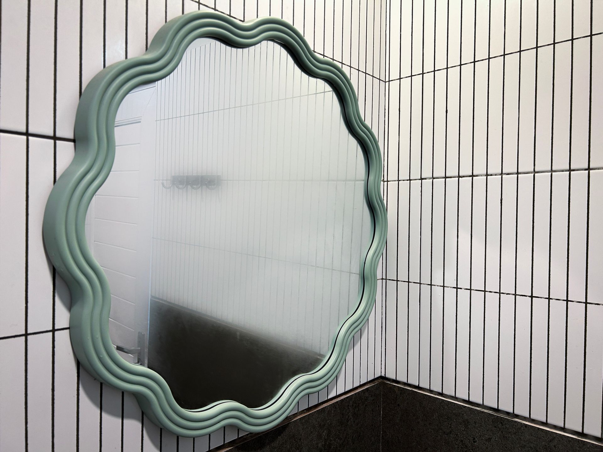 Wavy mint-green framed mirror on a white tiled wall above a dark countertop