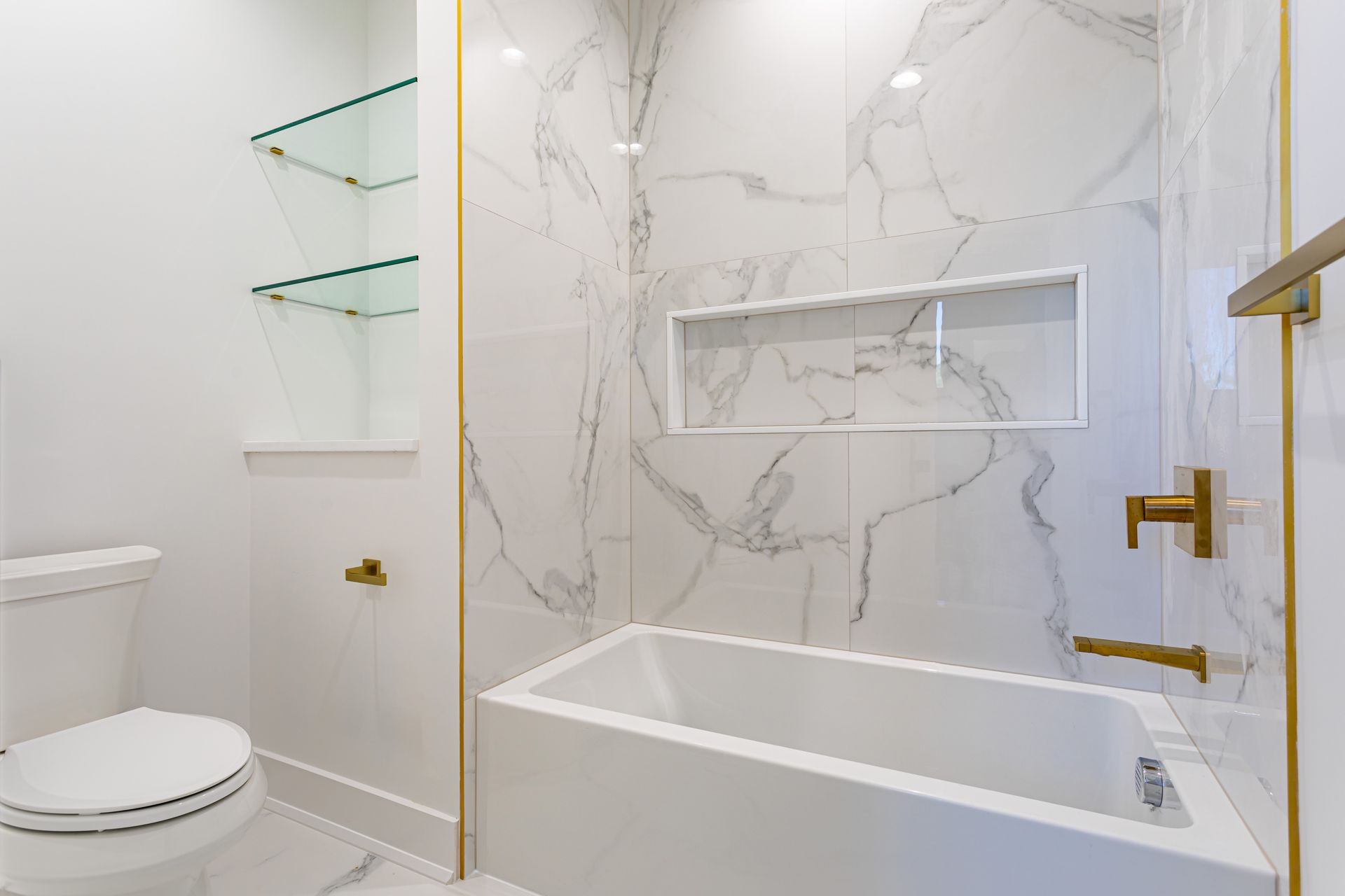 Modern white bathroom with marble tile, glass shelves, toilet, and a built-in bathtub.