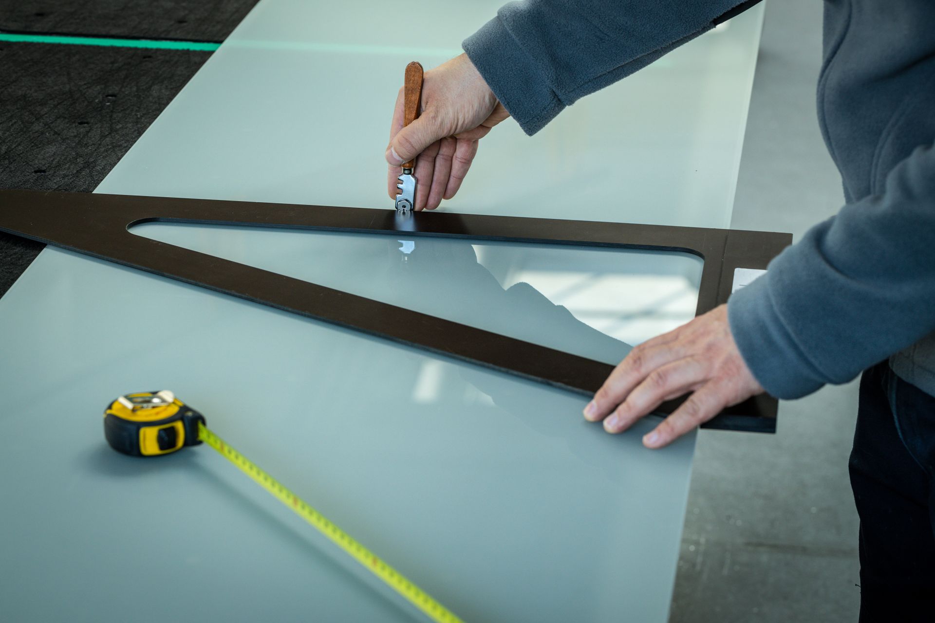 Person measuring and marking a glass tabletop with a tape measure and tool next to a metal frame