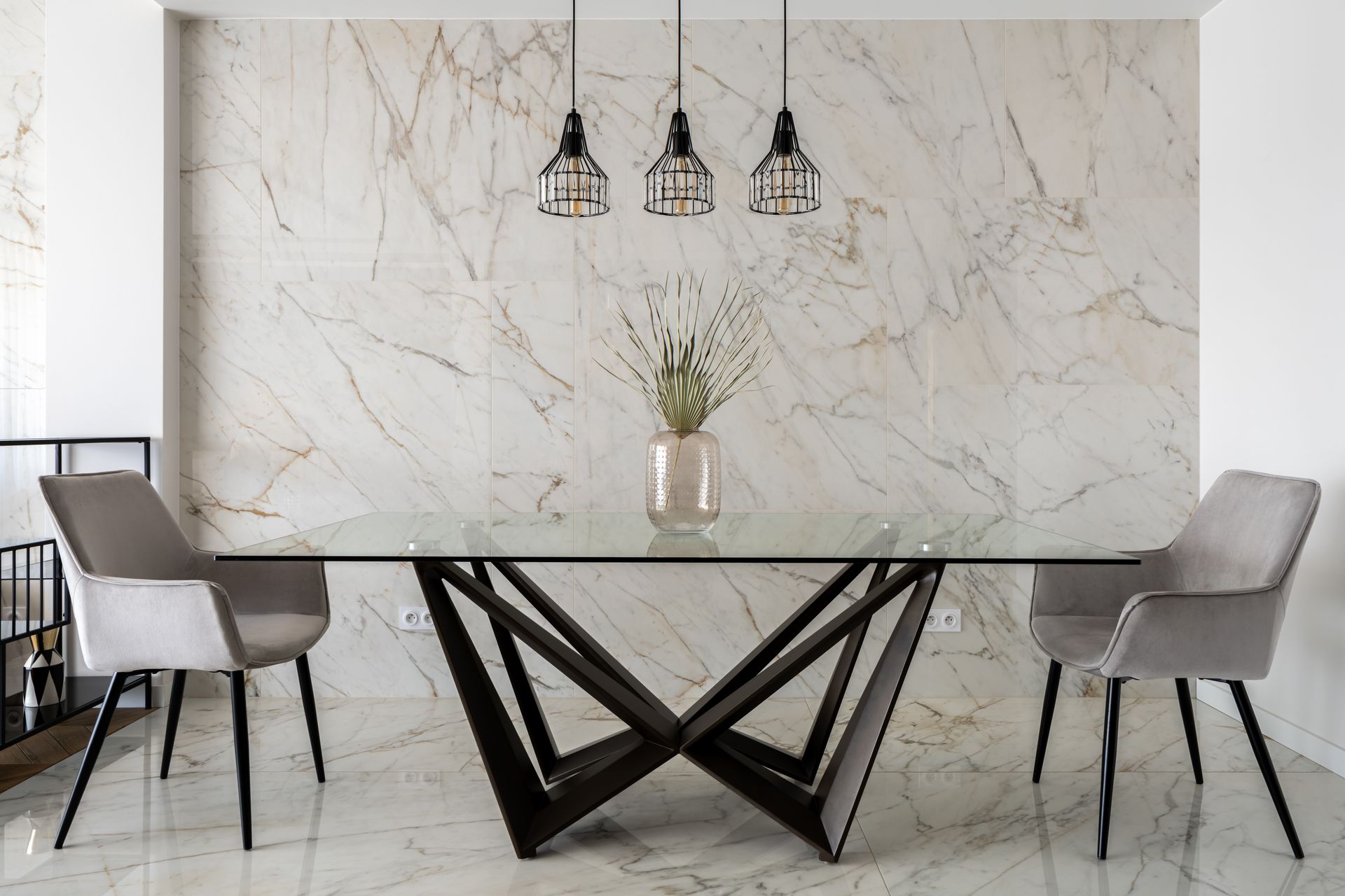 Modern dining room with marble wall, glass table, black geometric base, and gray chairs