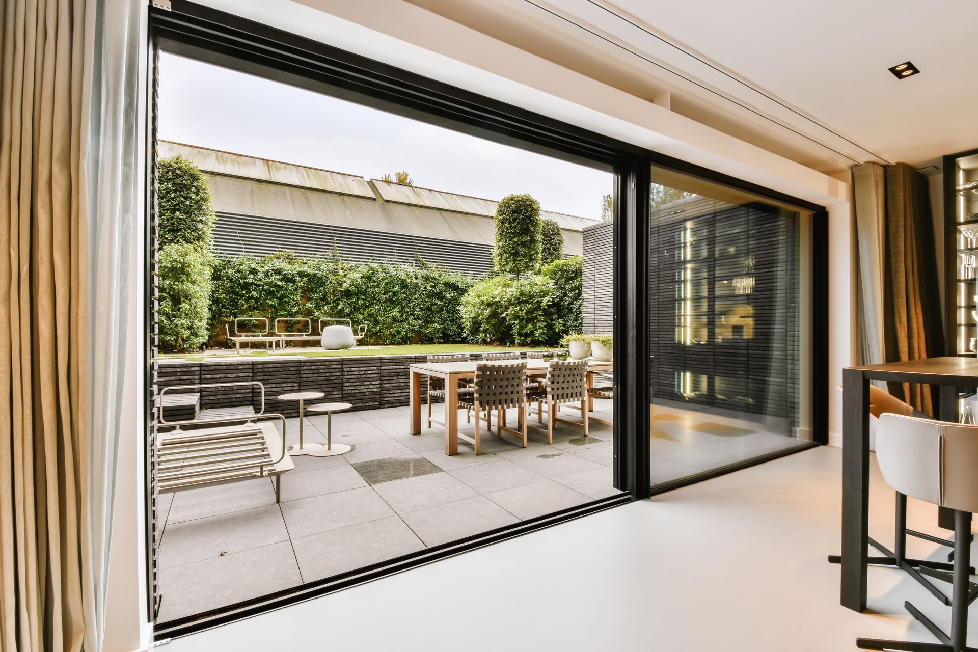 Modern patio seen through open sliding glass doors, with outdoor dining table and chairs beside a hedge-covered wall