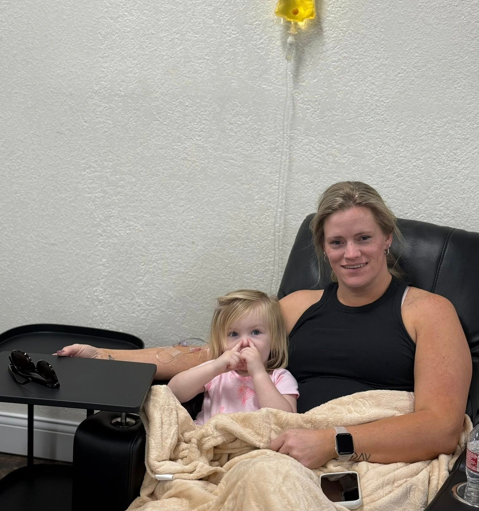 Woman and child sitting, woman receiving IV drip. Both wrapped in a blanket, smiling.
