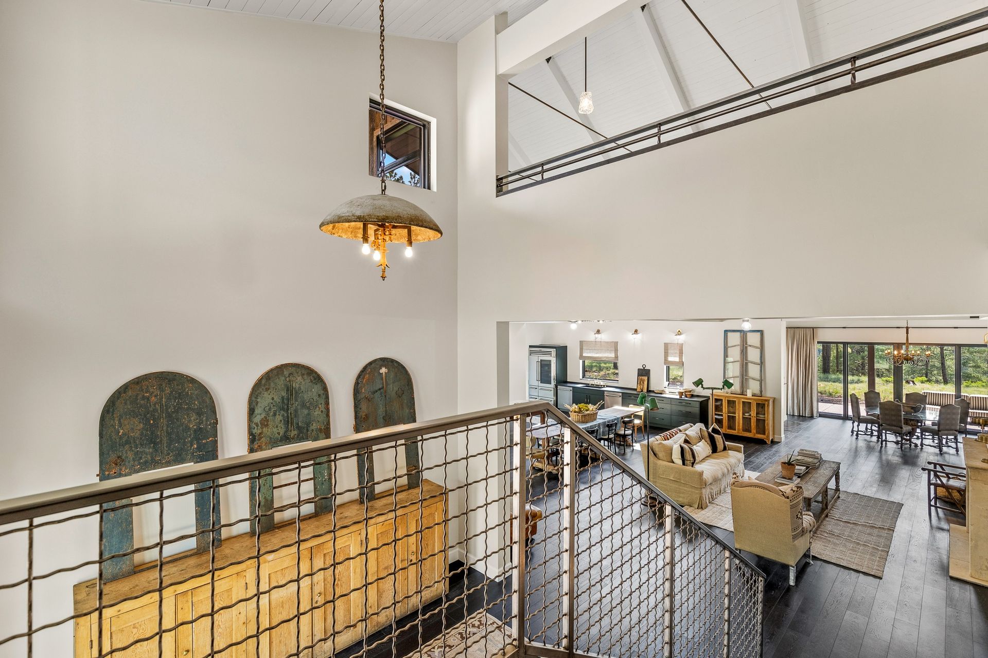 Bright loft-style office with mezzanine railing, hanging lights, desks, and large windows.