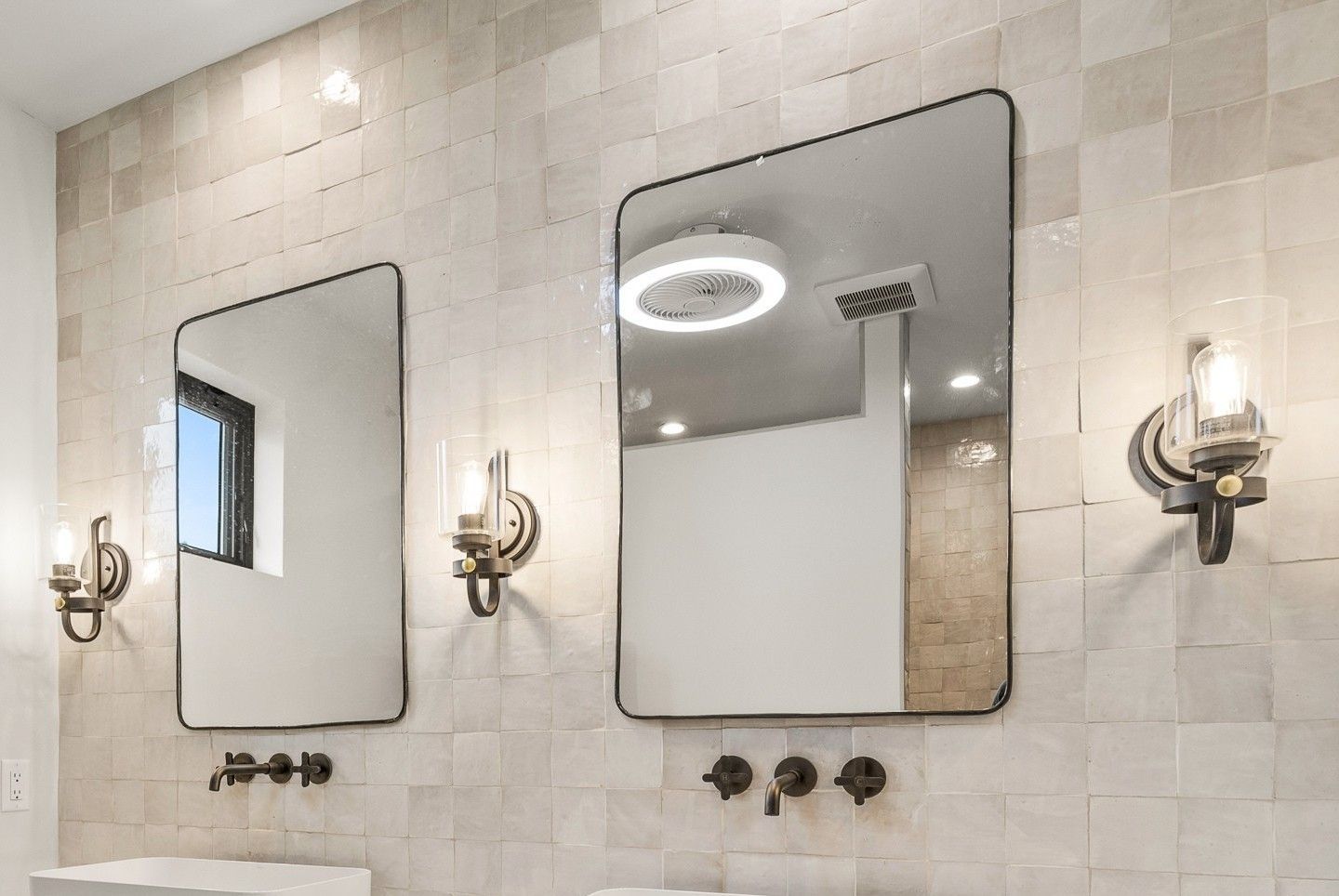 Bathroom mirrors above sinks on tiled wall with brass sconces and a window reflection