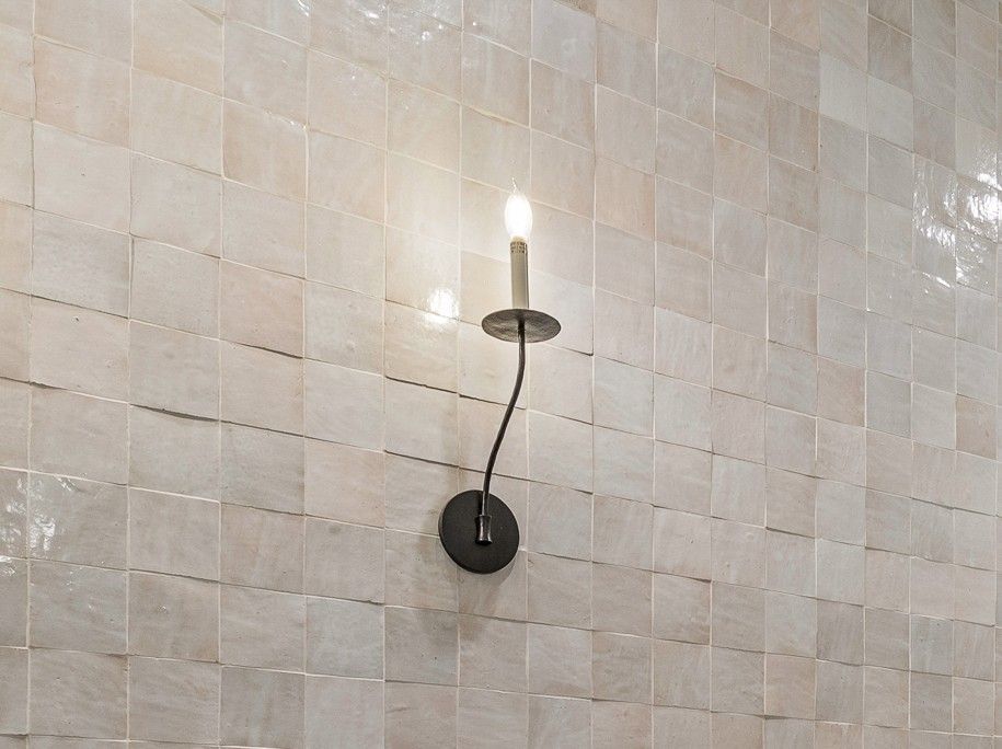 Wall-mounted candle sconce on a light tiled wall, with a small lit bulb above a curved black arm.