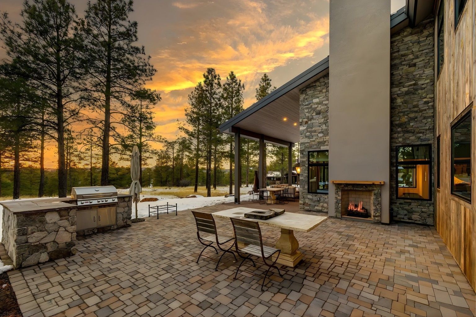 Patio with stone fireplace, table and chairs beside a modern cabin at sunset, with tall trees in the background