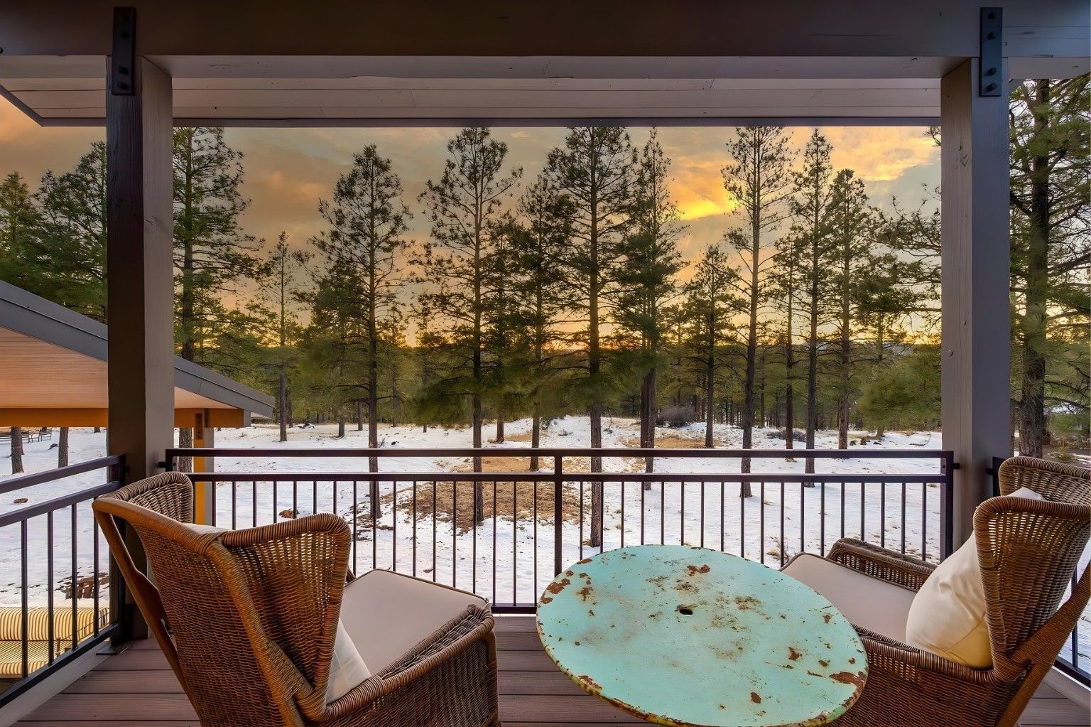 Snowy porch with wicker chairs and a turquoise table facing pine trees at sunset