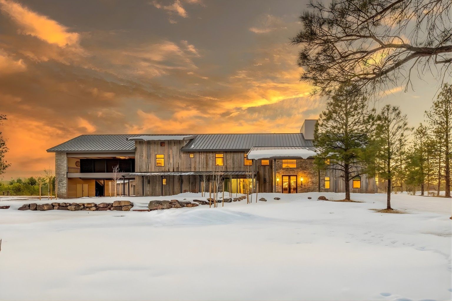Snow-covered modern cabin at sunset with warm lights glowing inside under a cloudy sky