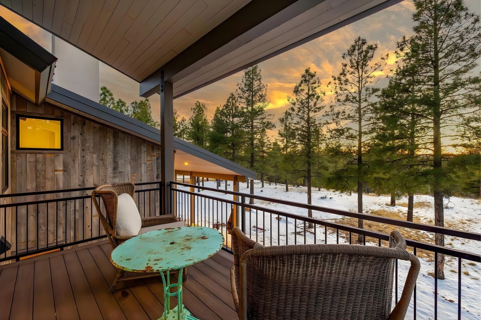 Covered balcony with wicker chairs and turquoise table overlooking snowy trees at sunset