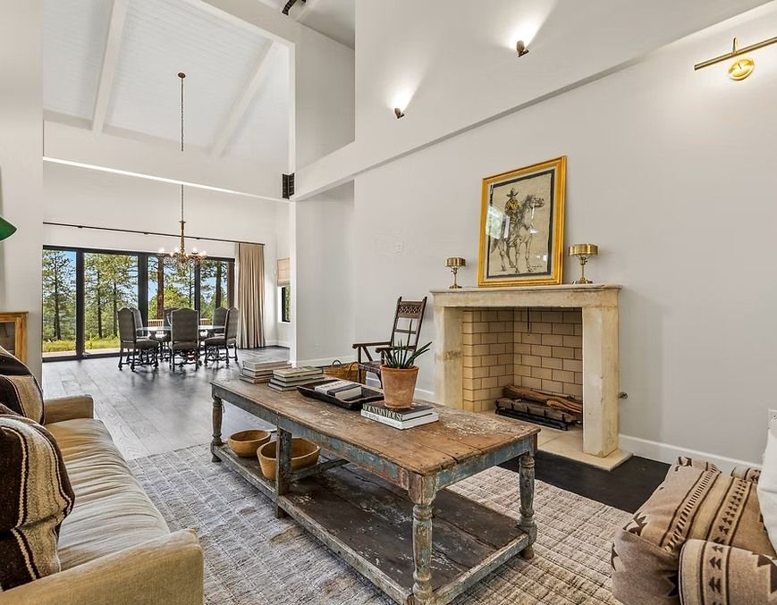 Bright living room with fireplace, high ceiling, large windows, and rustic coffee table