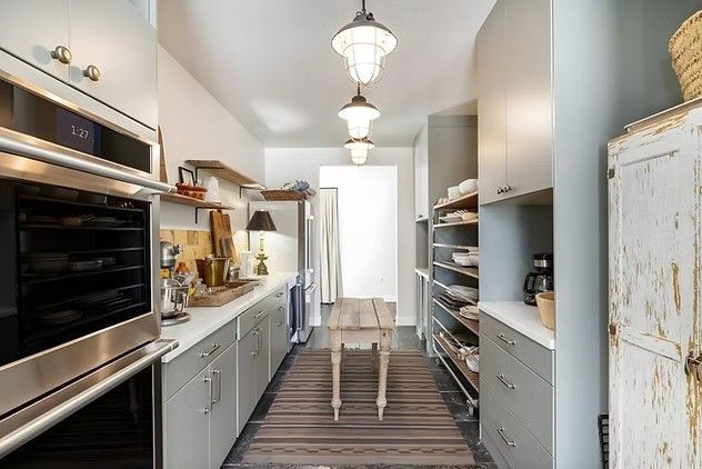 Narrow kitchen with white cabinetry, stainless oven, island table, and hanging pendant lights.
