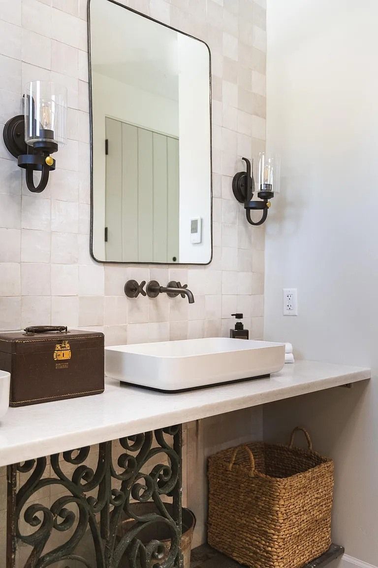 Bathroom vanity with white vessel sink, mirror, wall sconces, woven basket, and decorative ornate shelf