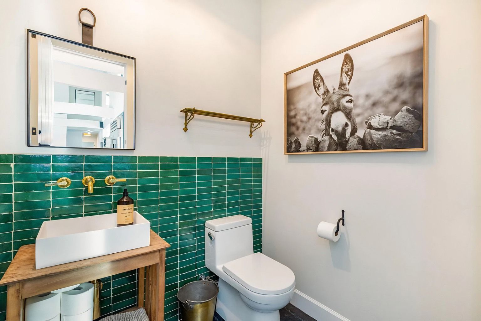 Compact bathroom with green tile, wooden vanity, toilet, mirror, and framed donkey artwork.