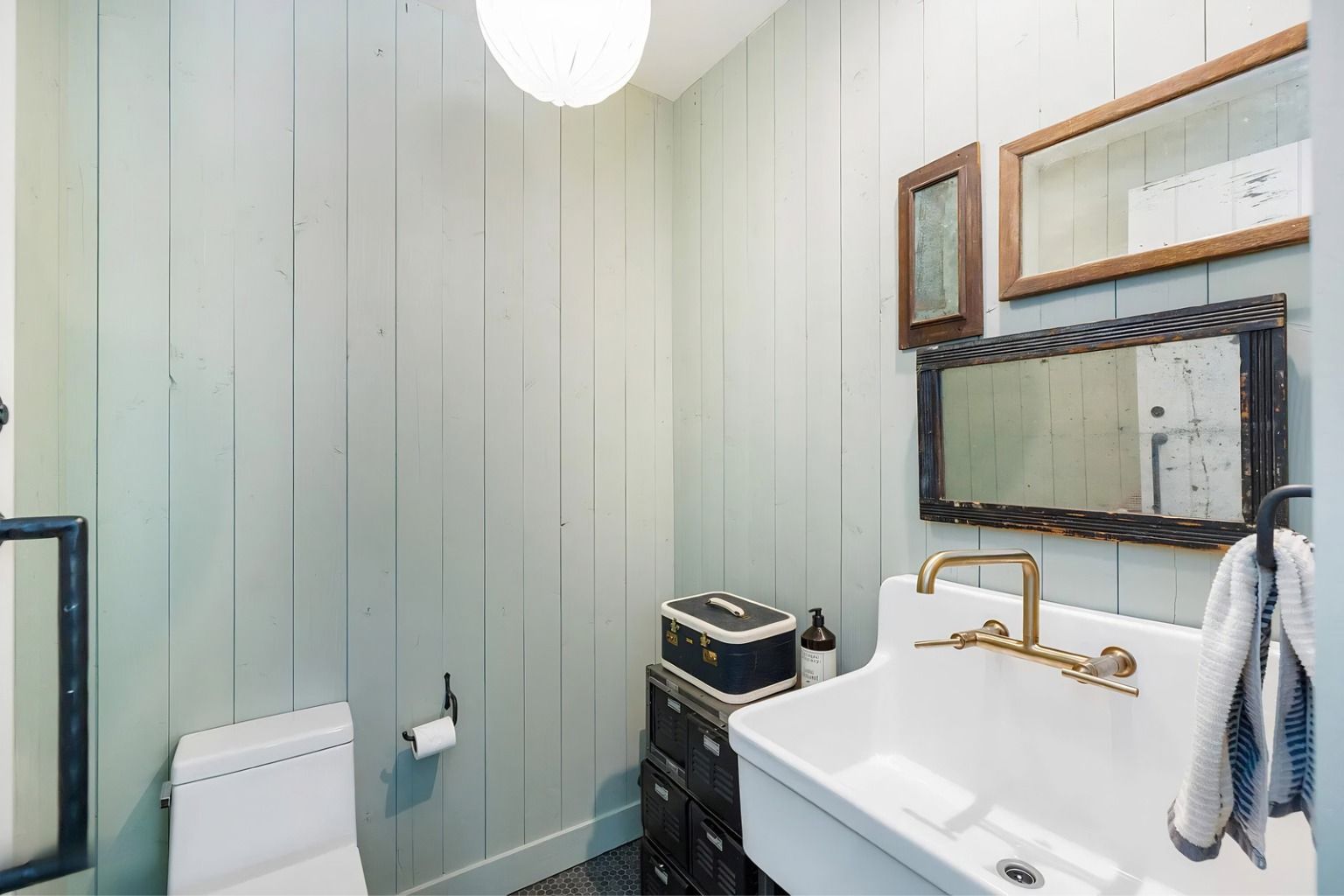 Small bathroom with white vanity, brass faucet, mirror, toilet, and towel rack, lit by a ceiling globe lamp.