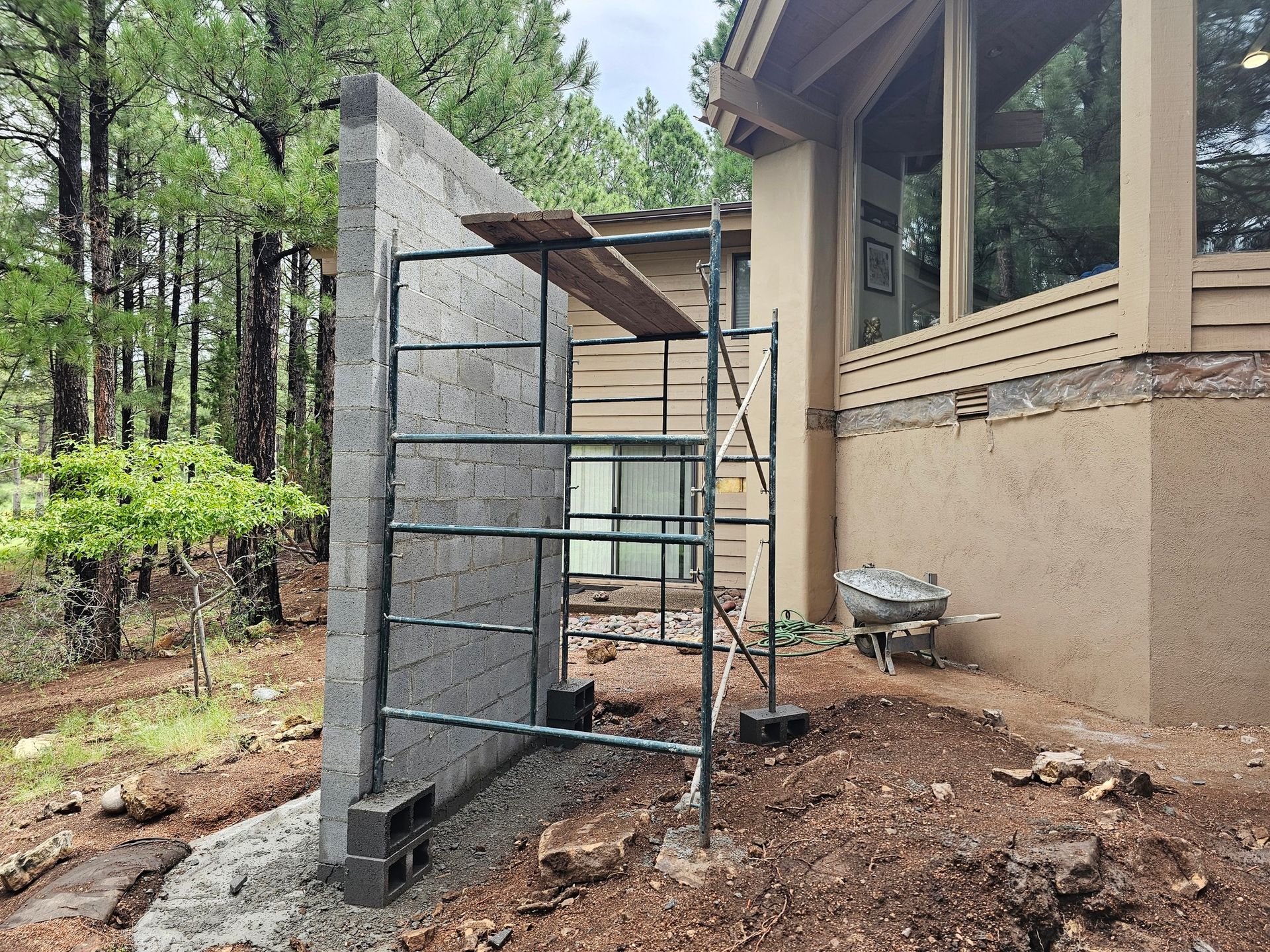 Scaffolding beside a house under construction, with a concrete wall and wooded yard.