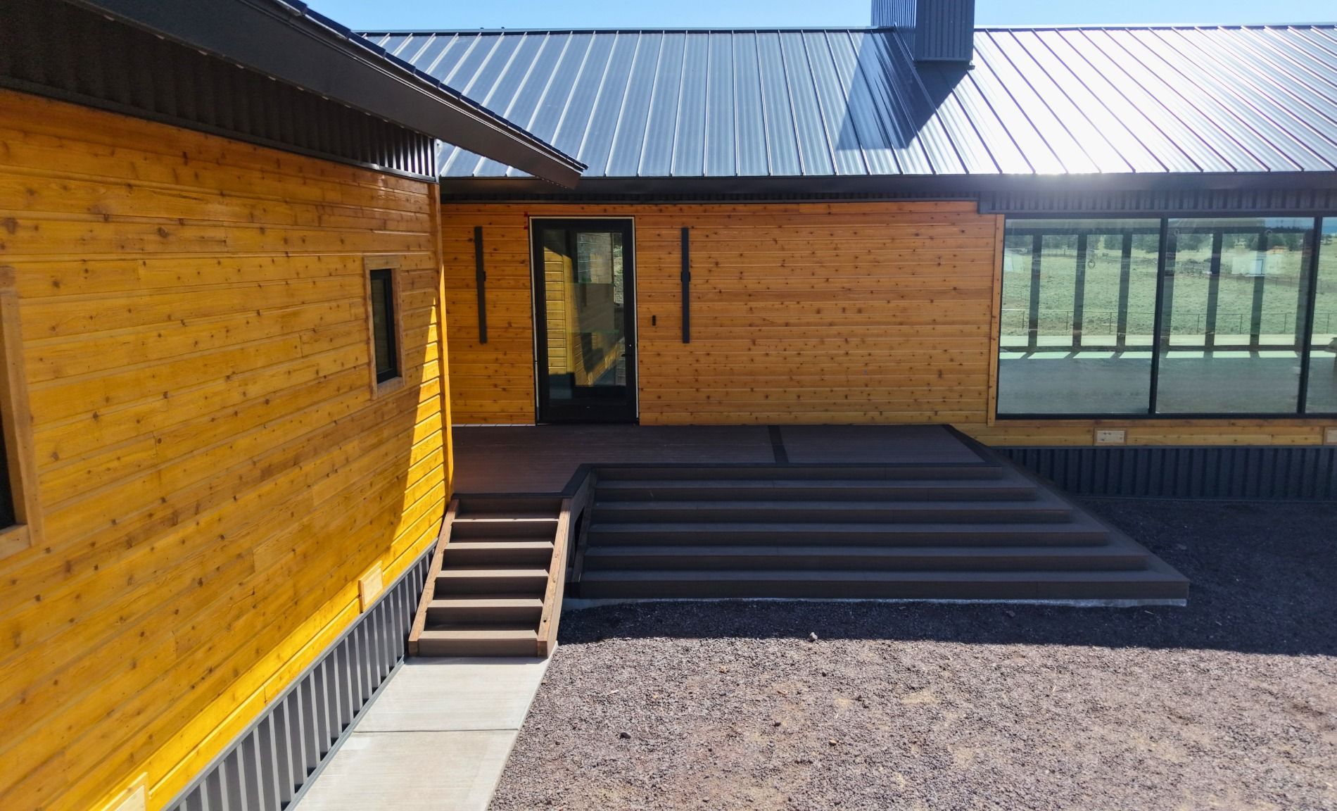 Modern wooden building with a sloped metal roof, steps, and large windows in a gravel courtyard