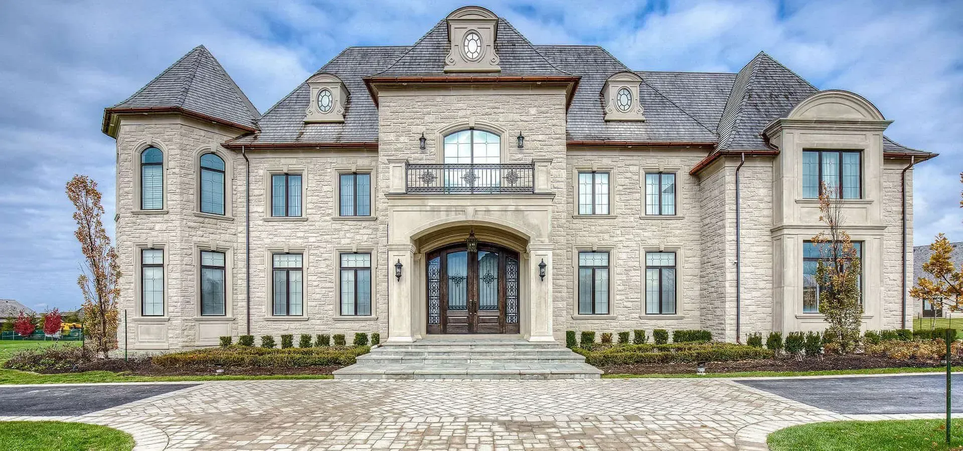 Large stone mansion with gray roof, arched entrance, and wide driveway under a cloudy sky