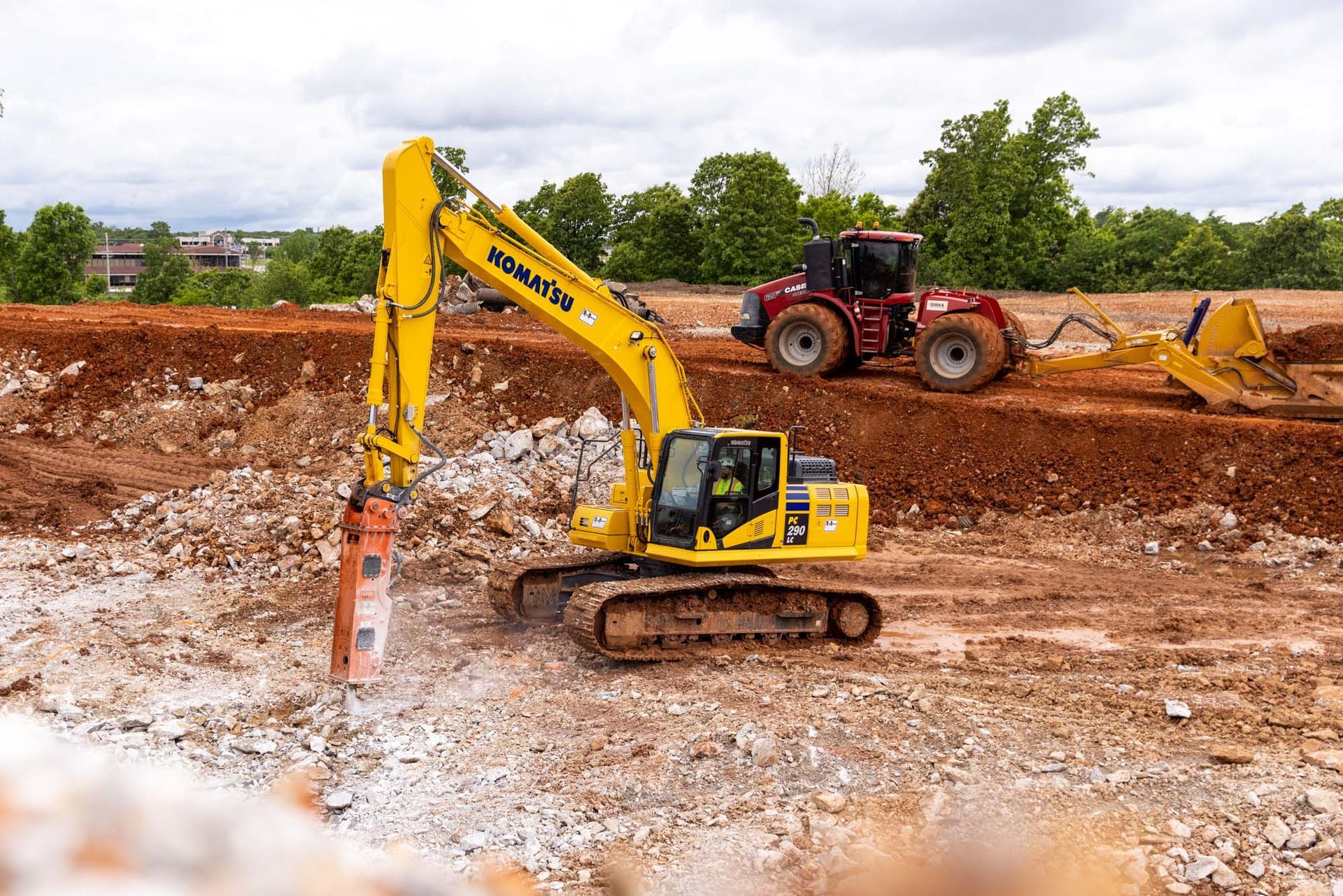 Yellow excavator digging in a muddy construction site, with a tractor and dirt pile in the background
