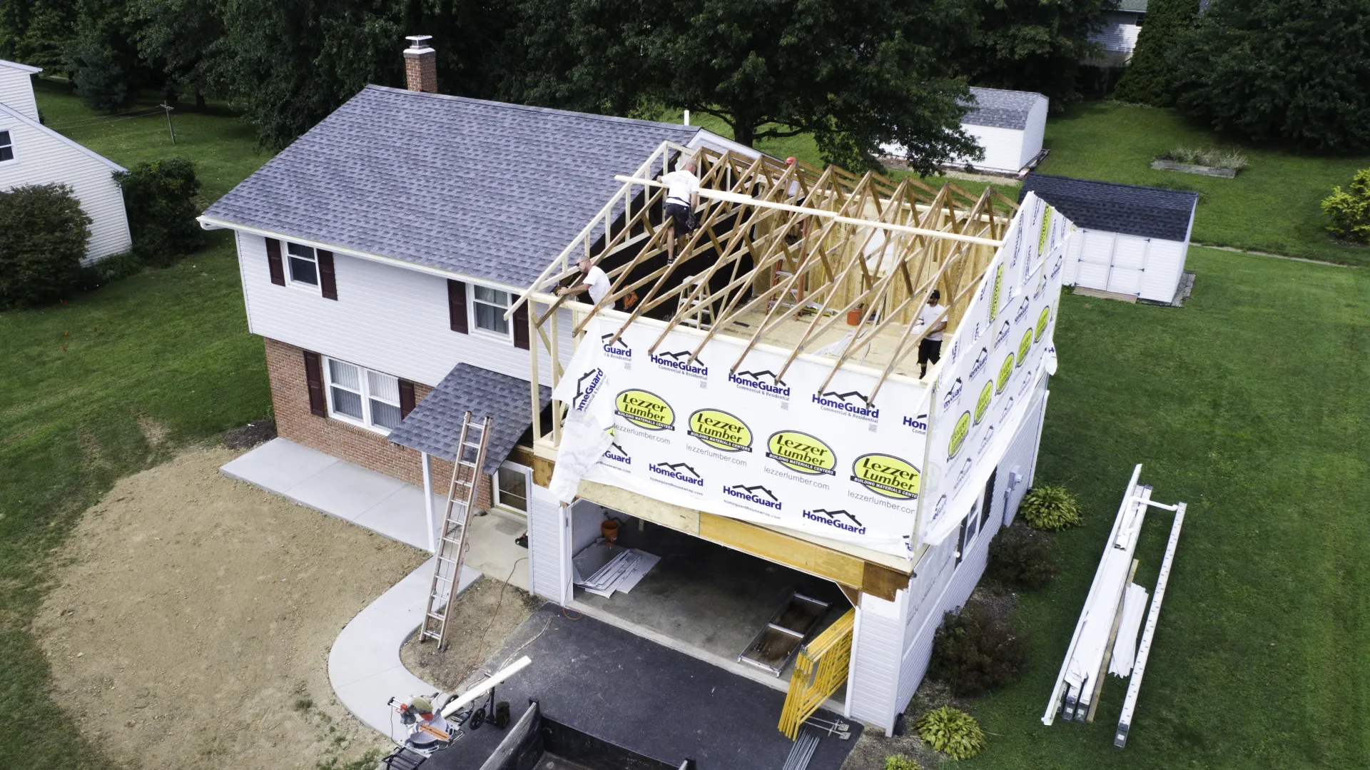 House under renovation with roof partially removed and exposed framing, white siding, and construction materials below