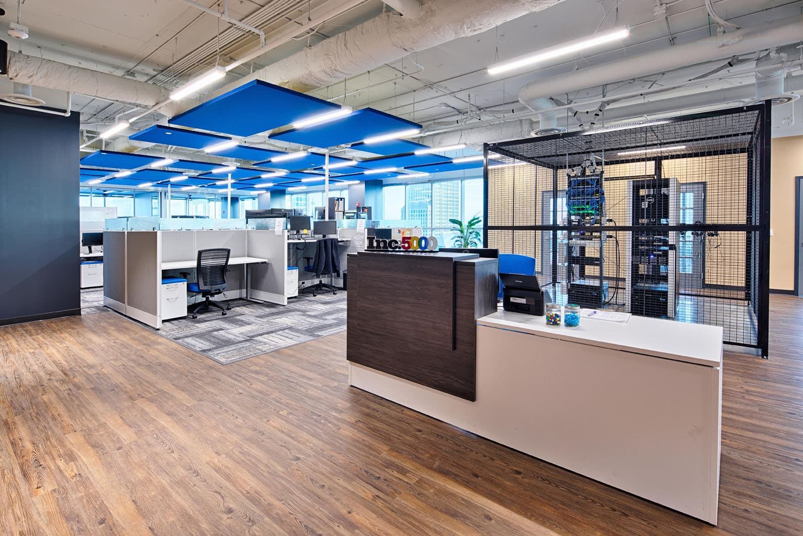 Modern open-plan office with desks, computers, blue ceiling panels, and wood flooring