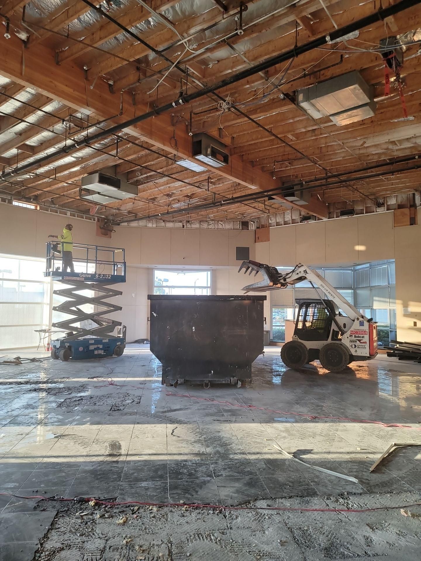 Warehouse interior with a scissor lift, skid steer, and dumpster under exposed ceiling framing.
