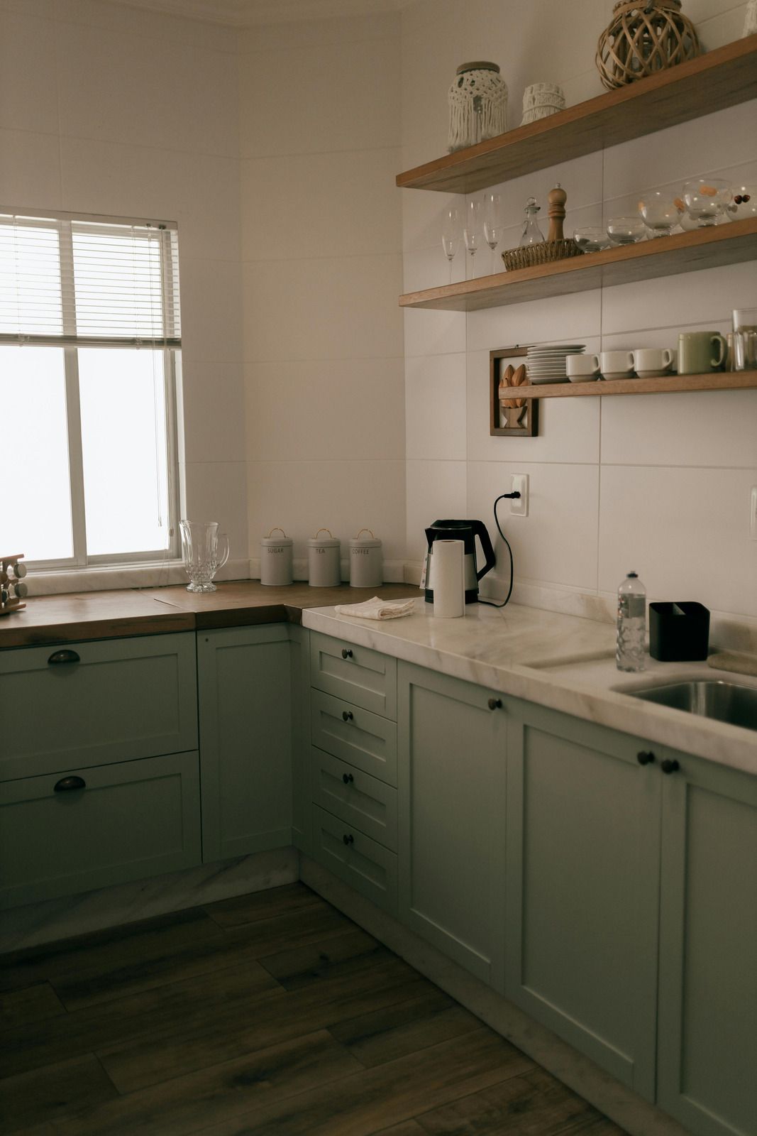 White kitchen with sage cabinets, wooden shelves, window, countertop items, and coffee machine