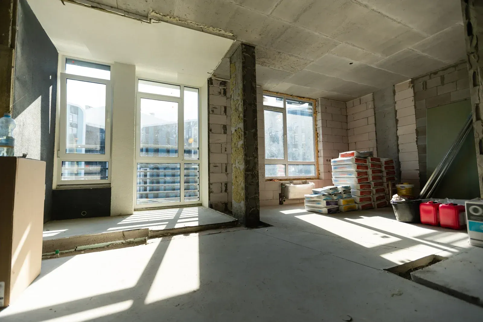 Unfinished apartment interior with large windows, exposed walls, and stacks of building materials in sunlight