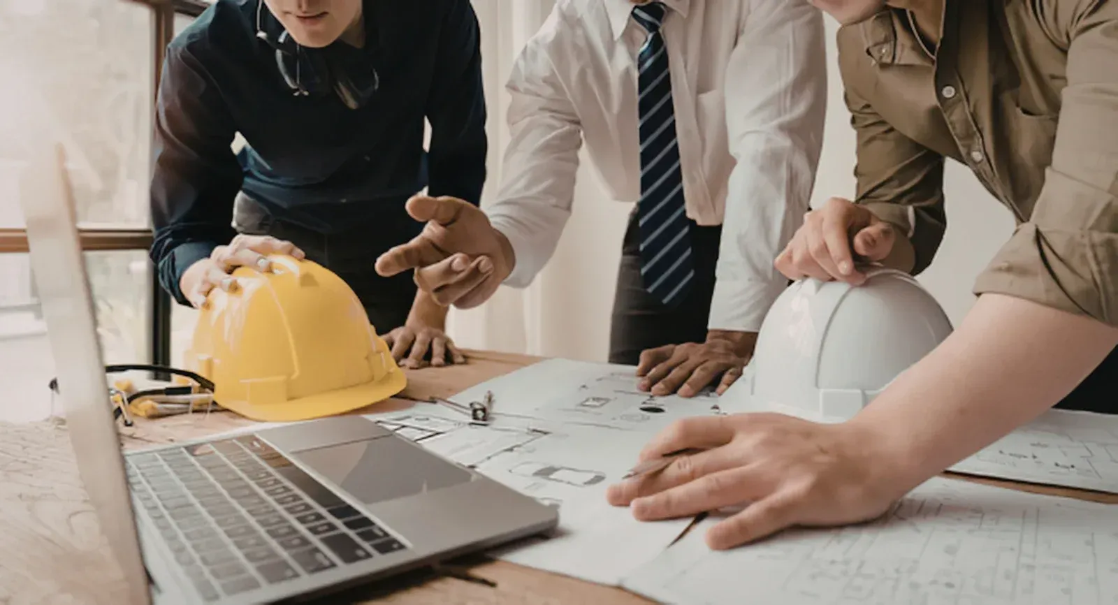 Colleagues reviewing blueprints around a laptop, yellow hard hat on a construction planning table
