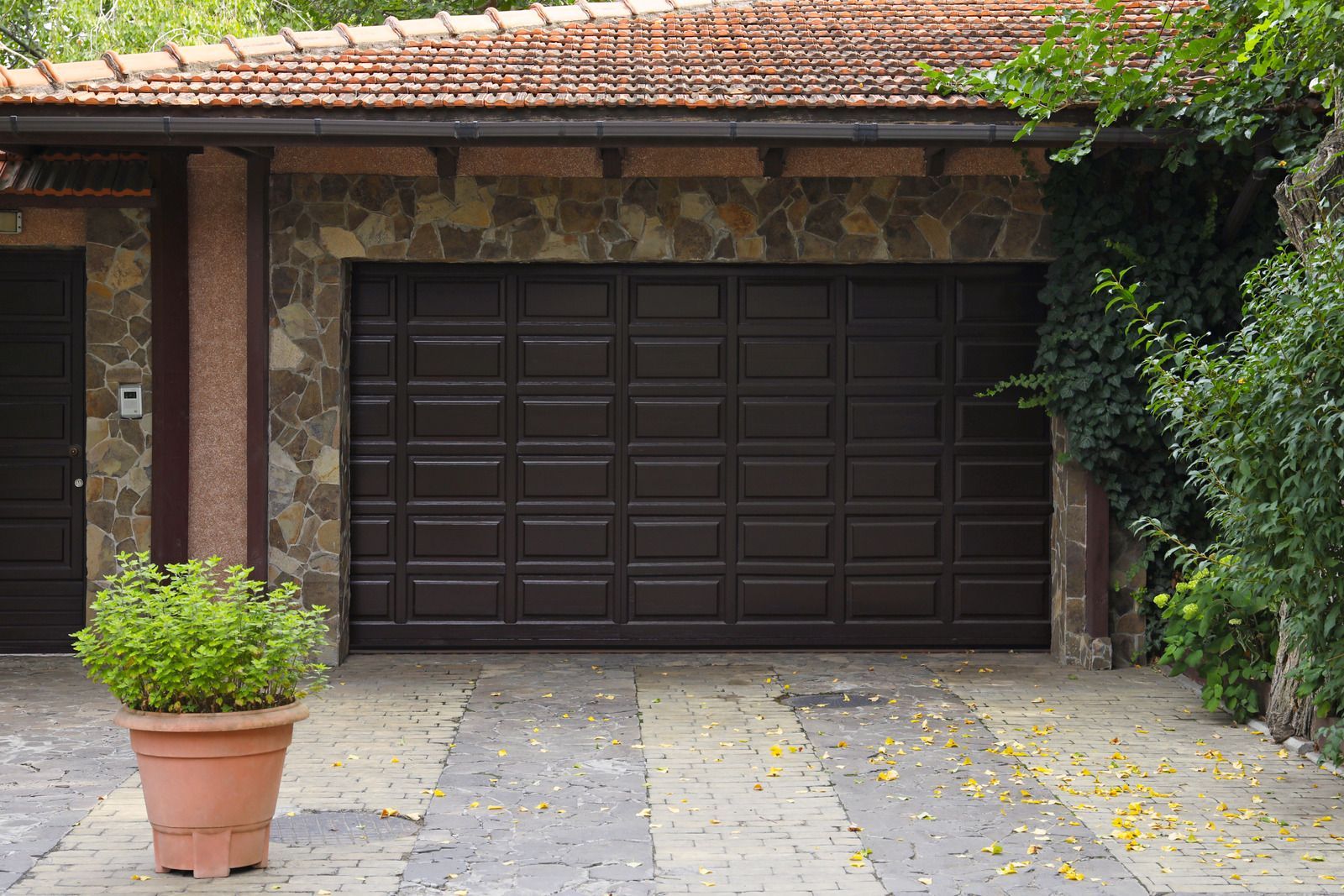 Stone house facade with a dark garage door, potted plant, and paved driveway