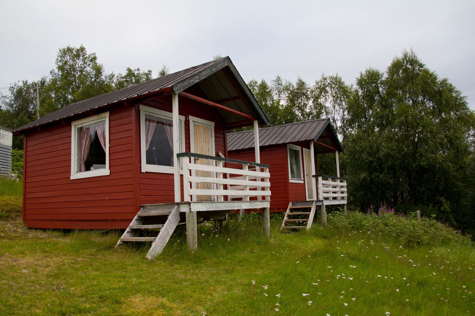 Two red cabins with white trim on a grassy hillside under cloudy sky