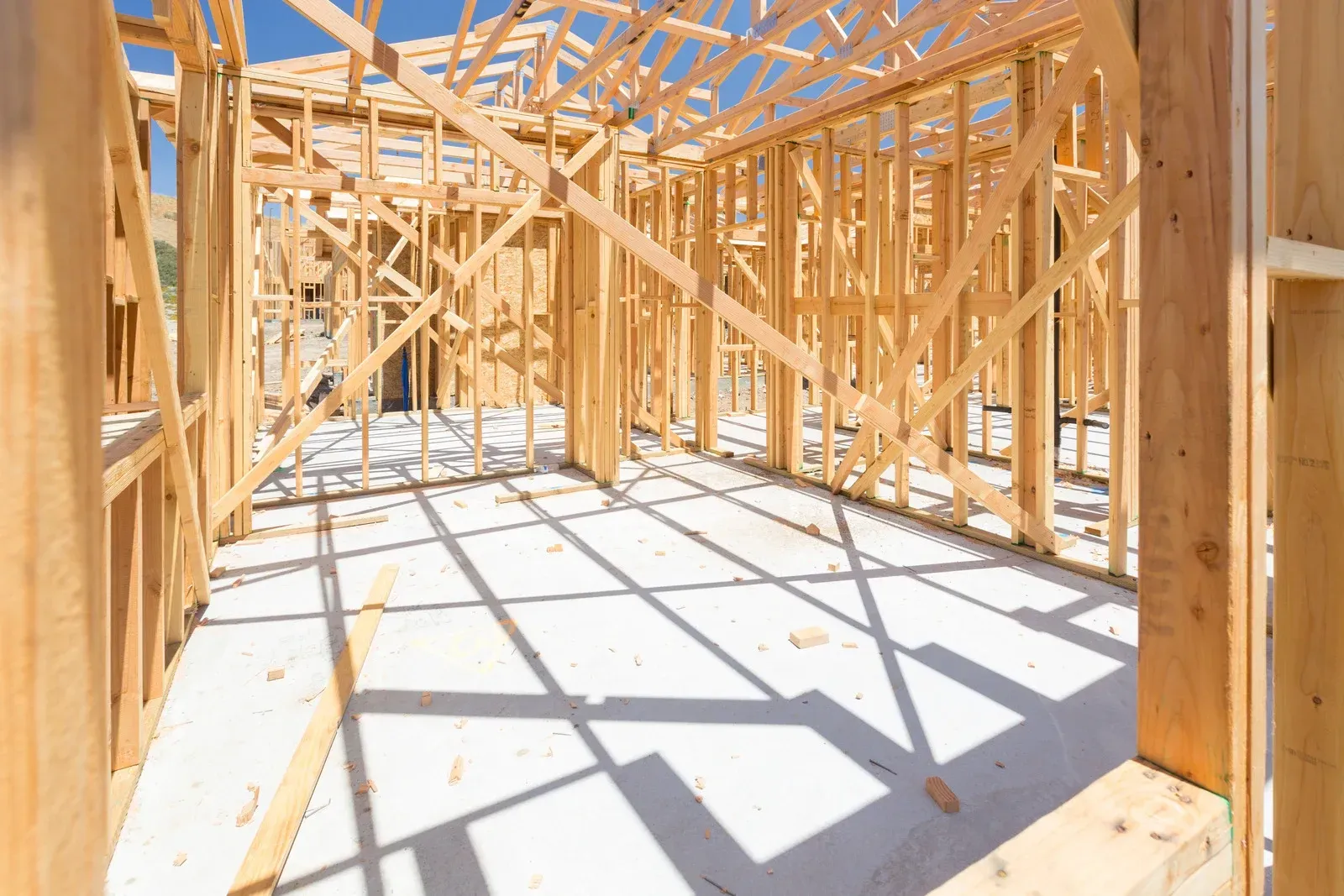 Interior of a house under construction with wooden framing and bright sunlight casting shadows on the concrete floor