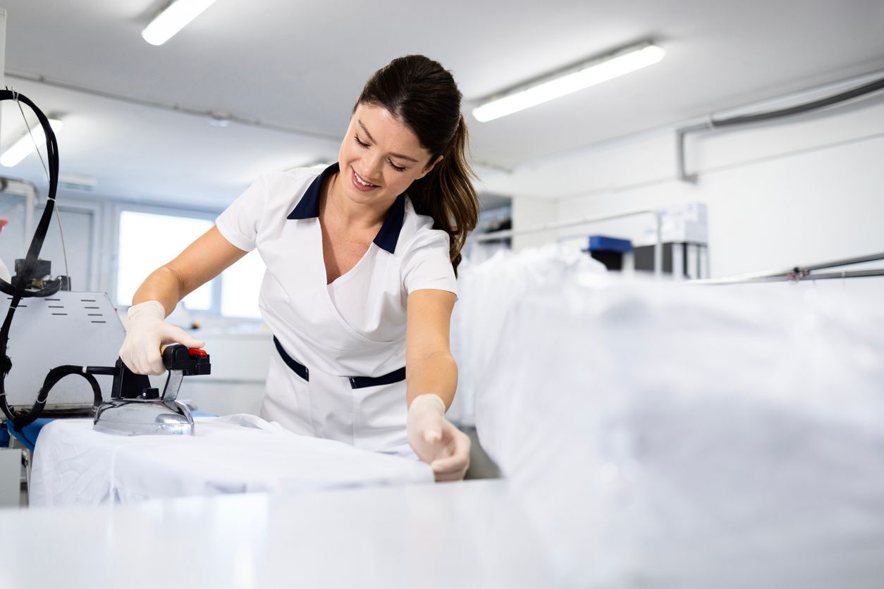 Woman in uniform ironing white fabric in a laundry room, smiling.