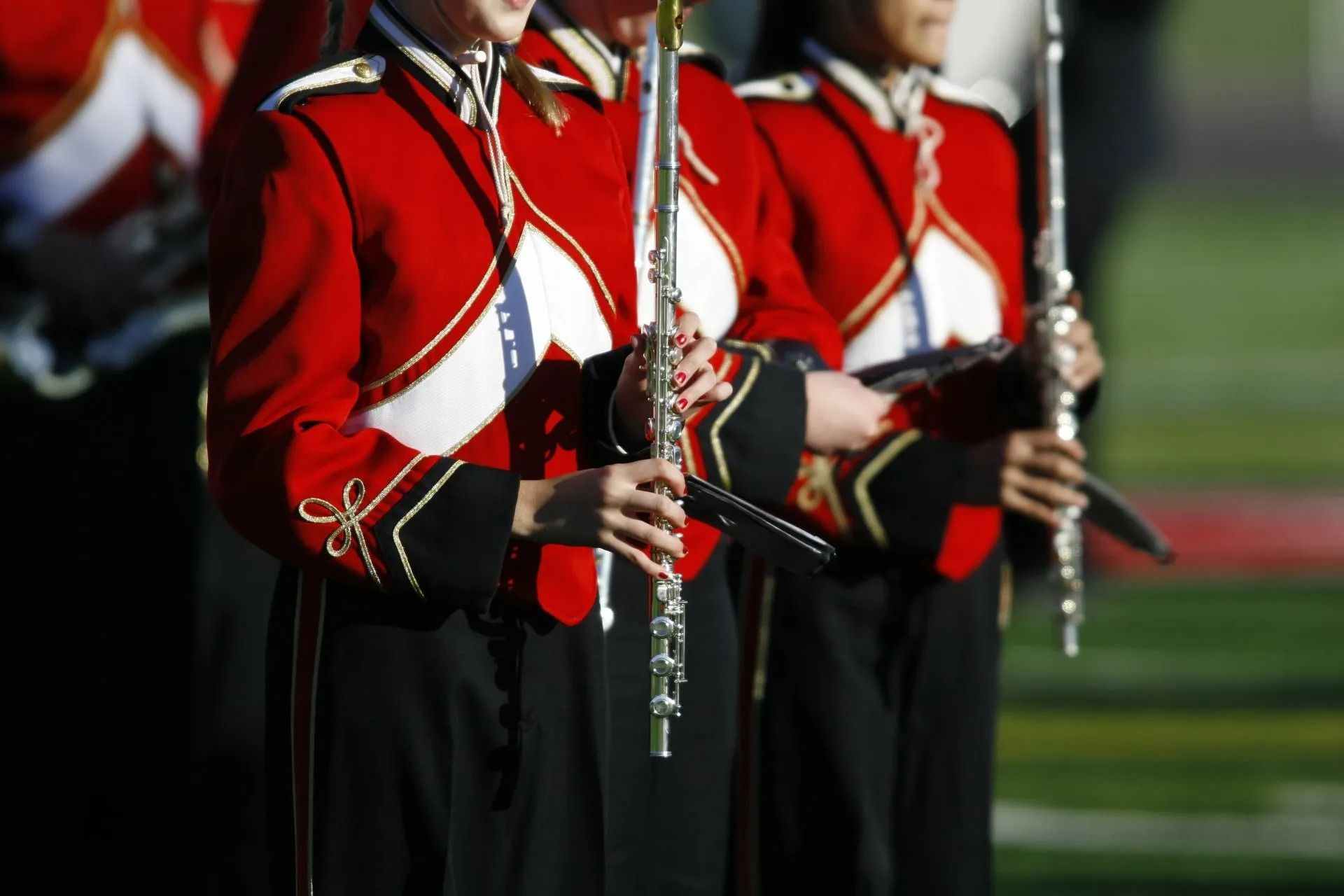 Marching band members in red jackets with flutes on a green field.
