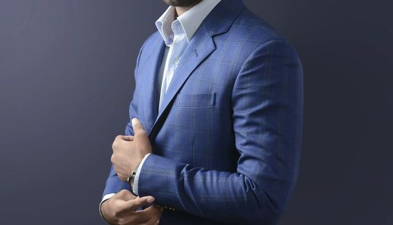 Man in blue blazer and white shirt, hand on lapel, against a dark blue background.
