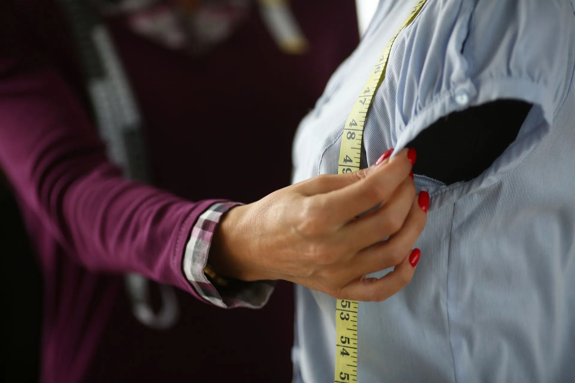 Person measuring sleeve of blue shirt on a mannequin with a measuring tape. Purple shirt in background.