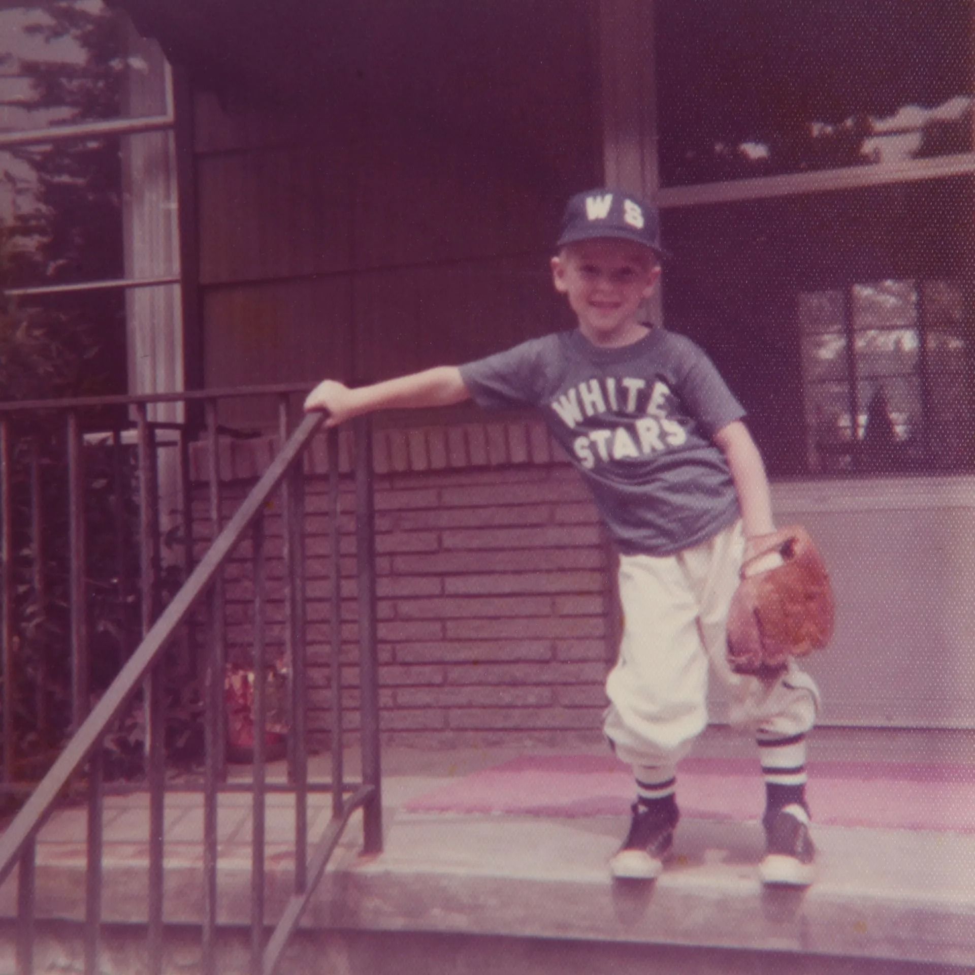 Young boy in baseball uniform holds glove, stands on porch.