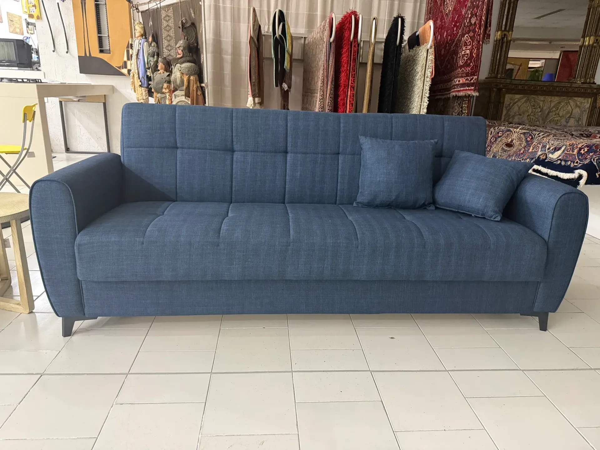 Divano imbottito blu con schienale capitonné e due cuscini coordinati su sfondo bianco.