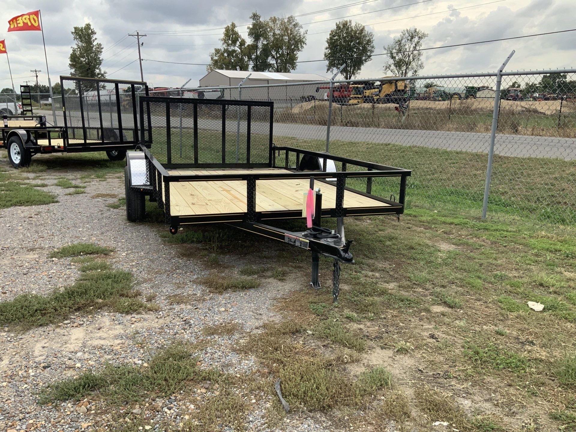 Black utility trailer with red jack on gravel beside a chain-link fence in a yard