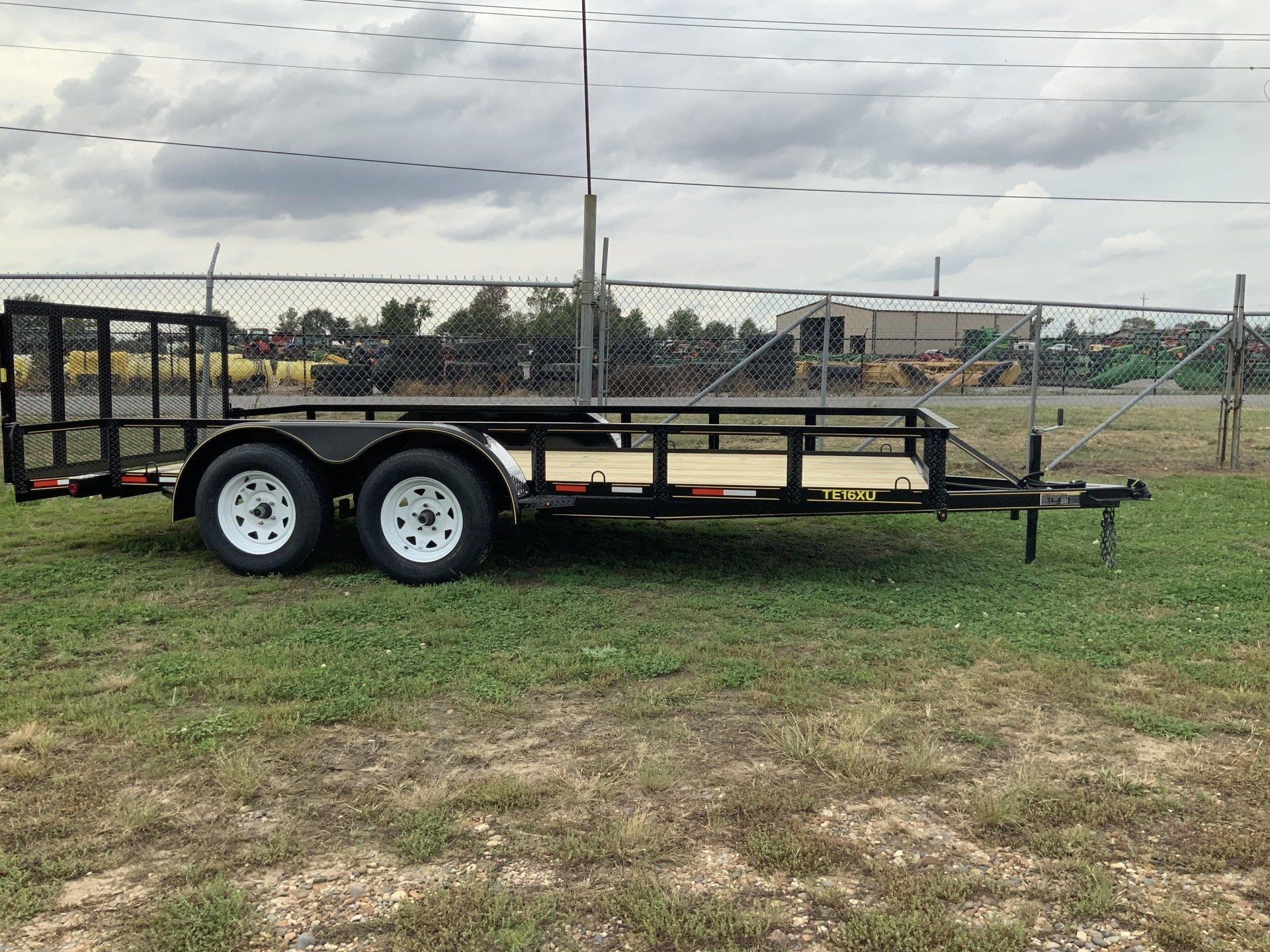 Black utility trailer with dual axles parked on grass under a cloudy sky.