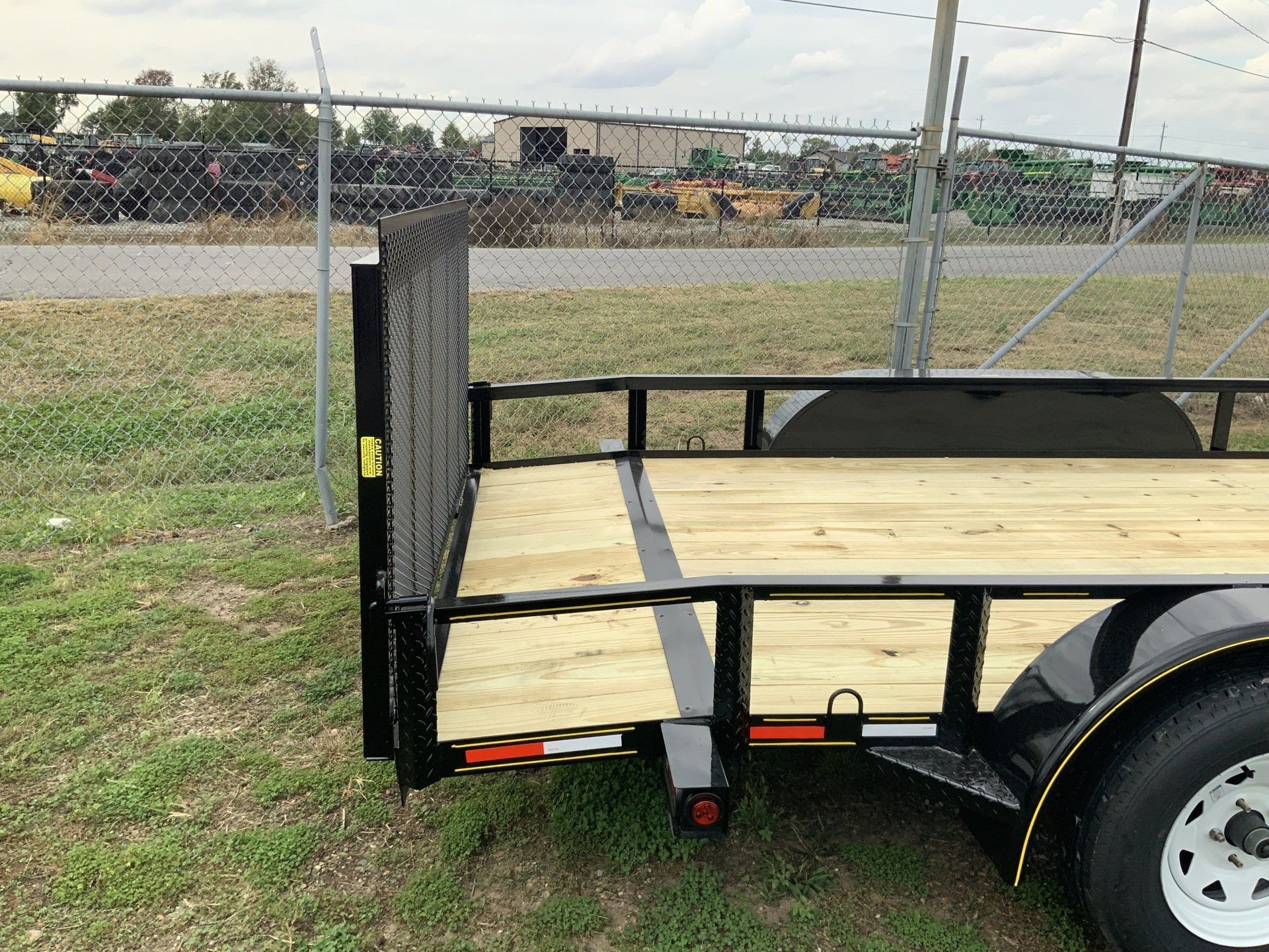 Small flatbed utility trailer with black metal frame and plywood floor, parked on grass near a chain-link fence.