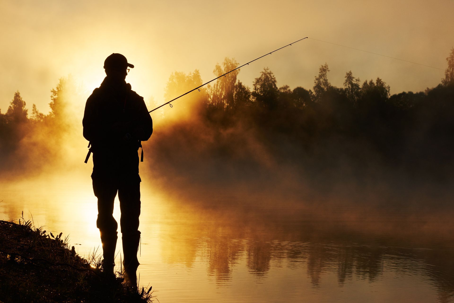 A man is fishing in a foggy lake at sunset