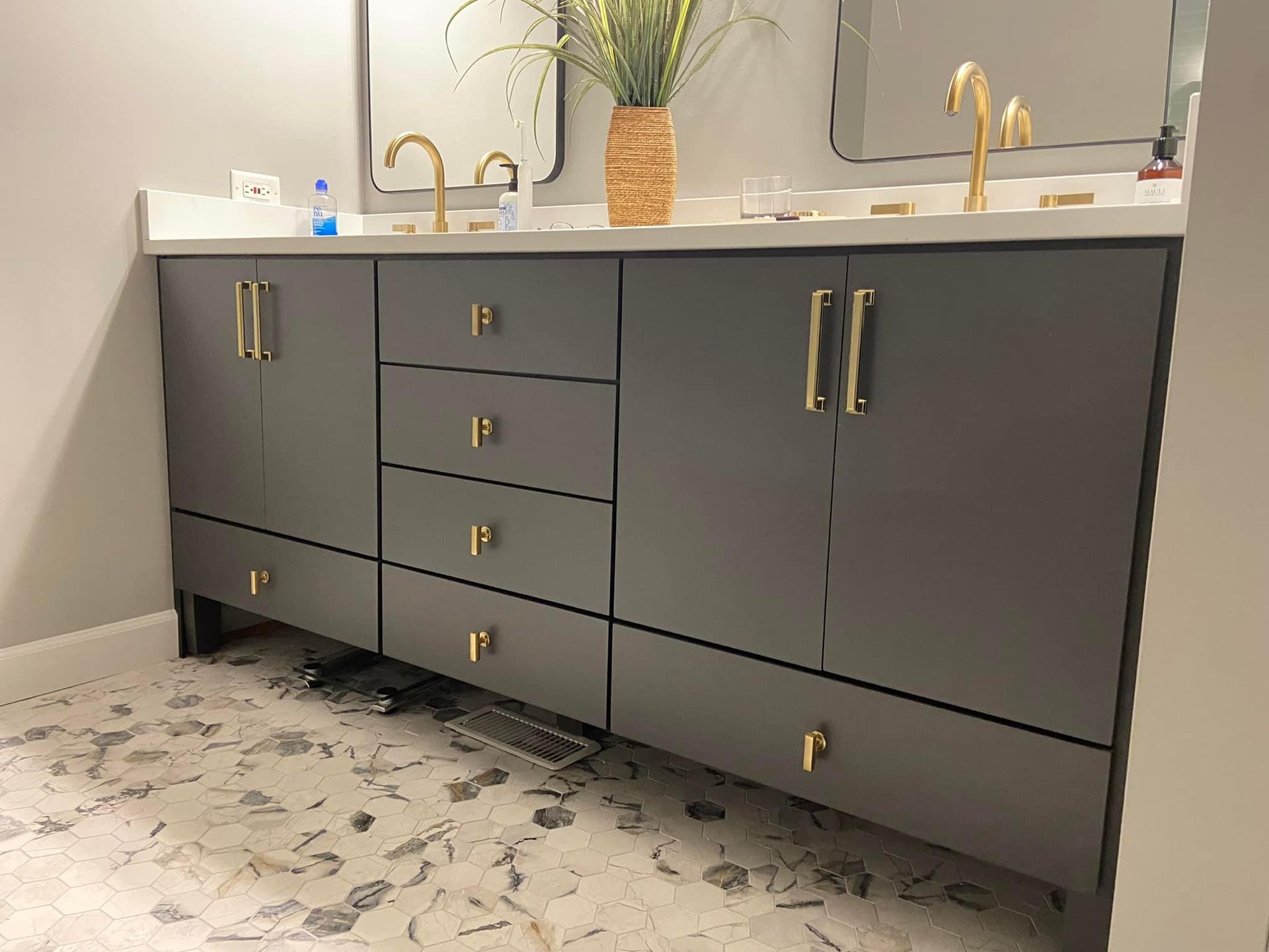 Modern gray bathroom vanity with gold hardware, white countertop, and dual gold faucets on patterned tile floor