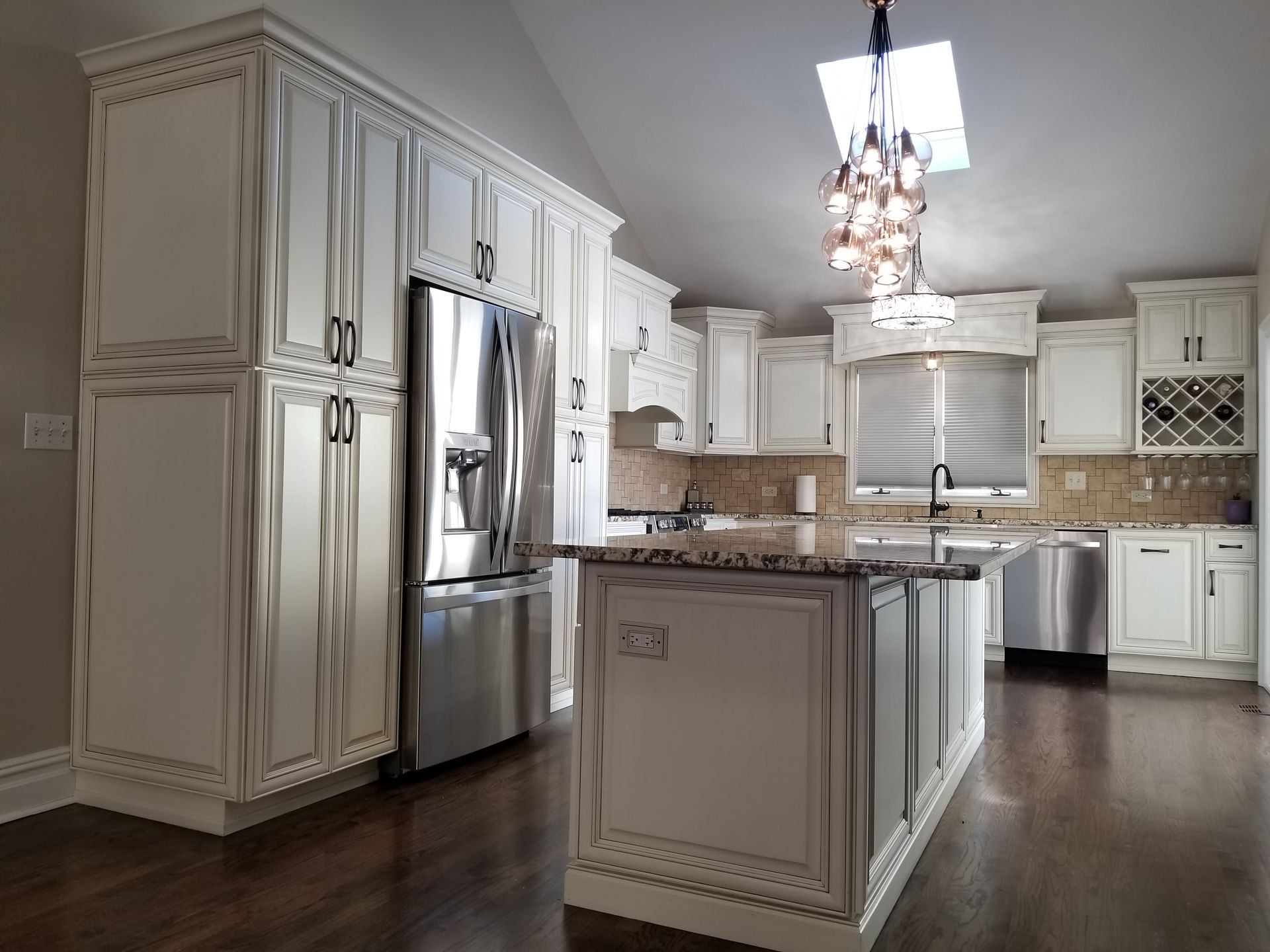 Bright white kitchen with stainless steel appliances, island, and chandelier under a skylight.