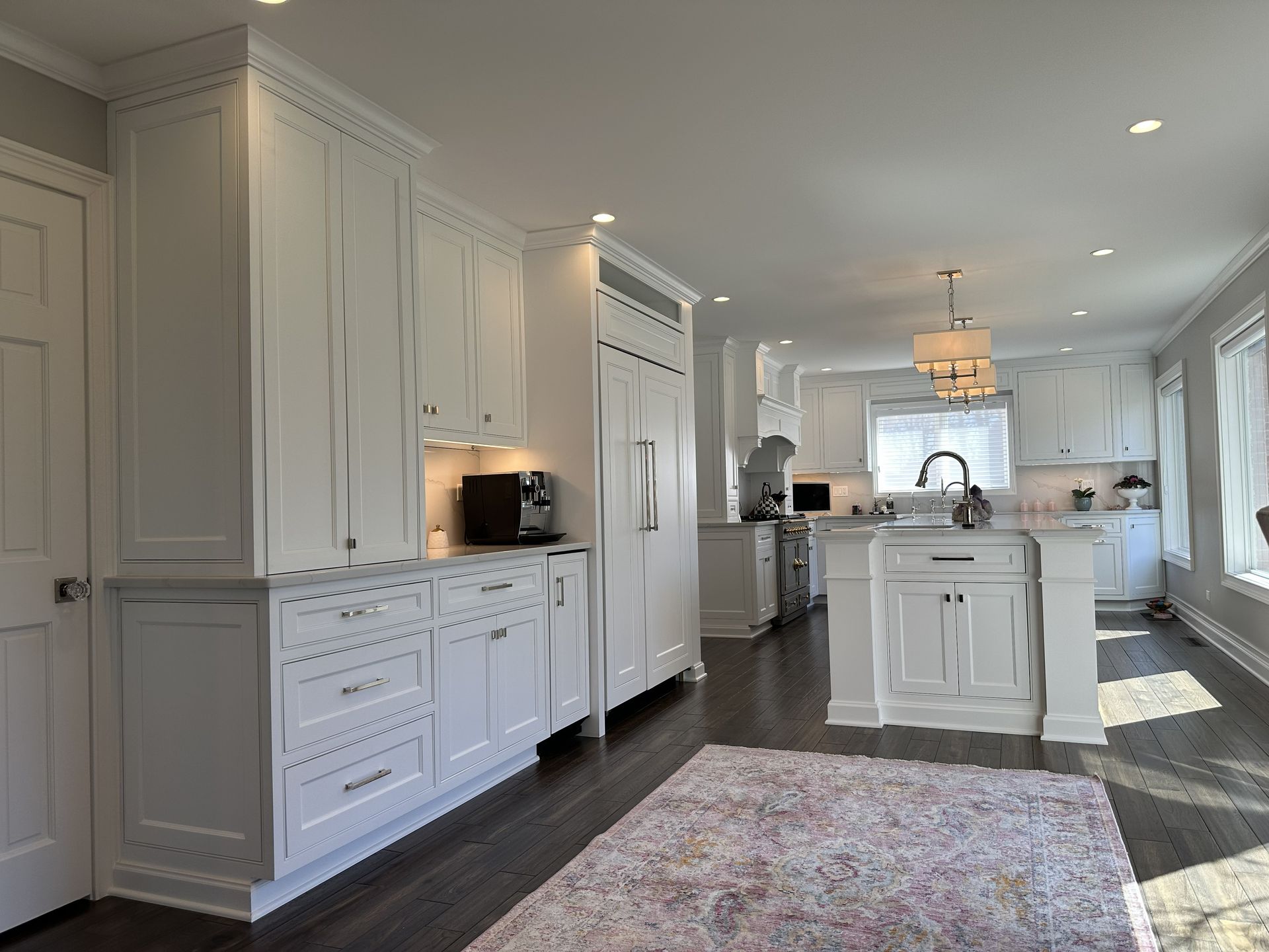 Bright white kitchen with island, tall cabinets, dark wood floors, and a patterned rug under recessed lights