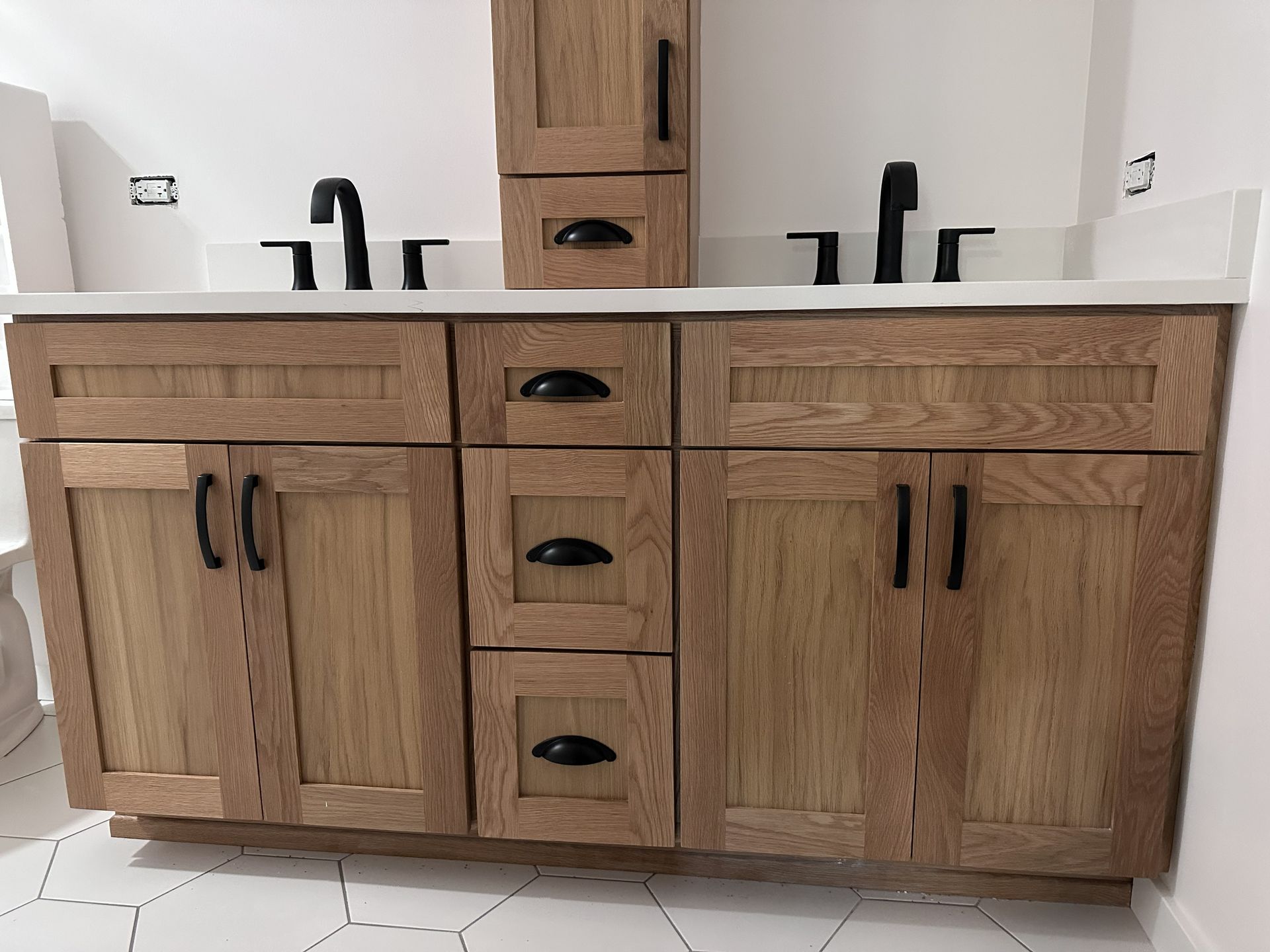 Wooden double vanity with white countertop, black fixtures, and a center drawer stack in a bathroom.