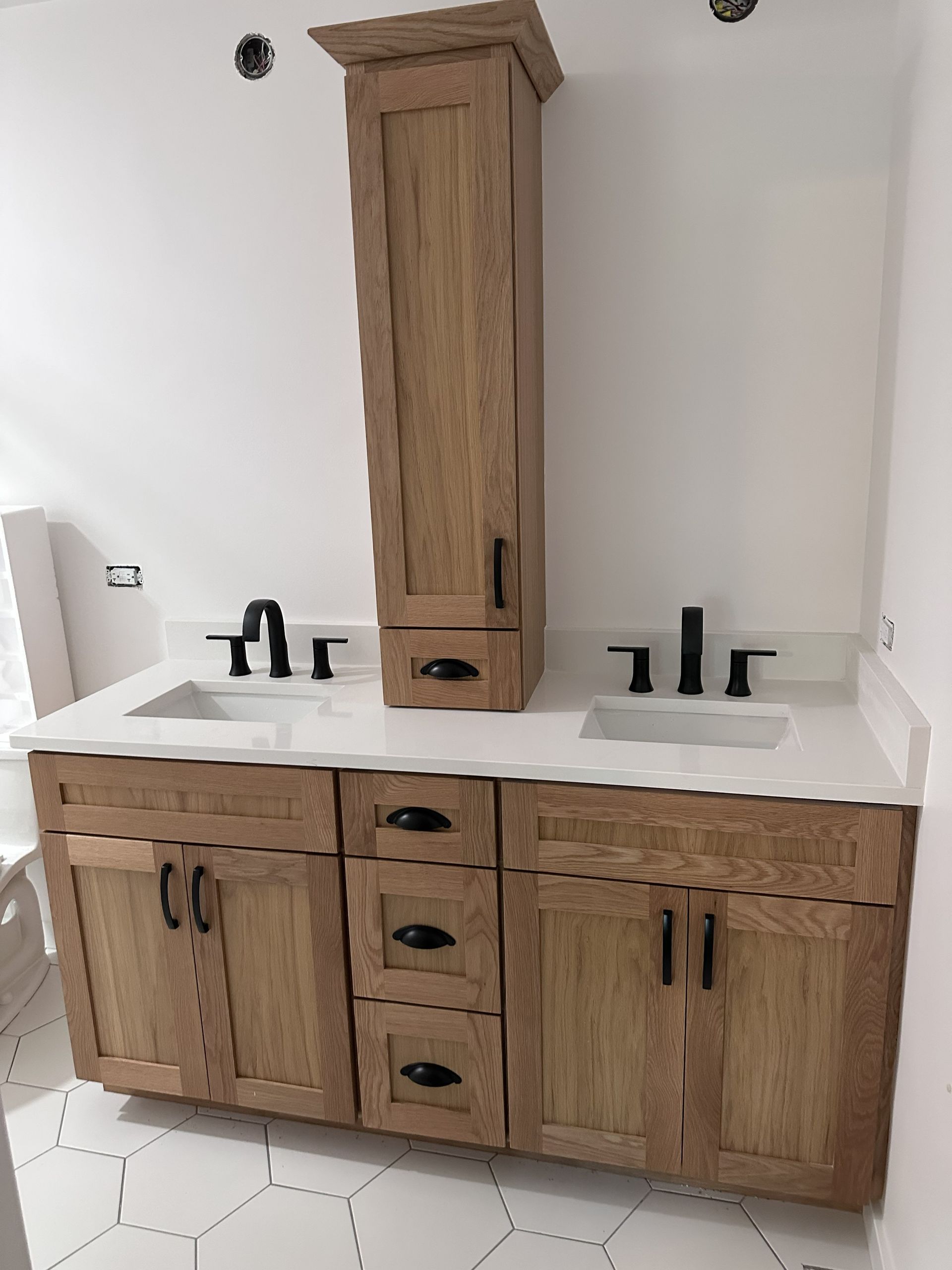 Wood bathroom vanity with double sinks, black fixtures, and a tall center cabinet on white hex tile floor