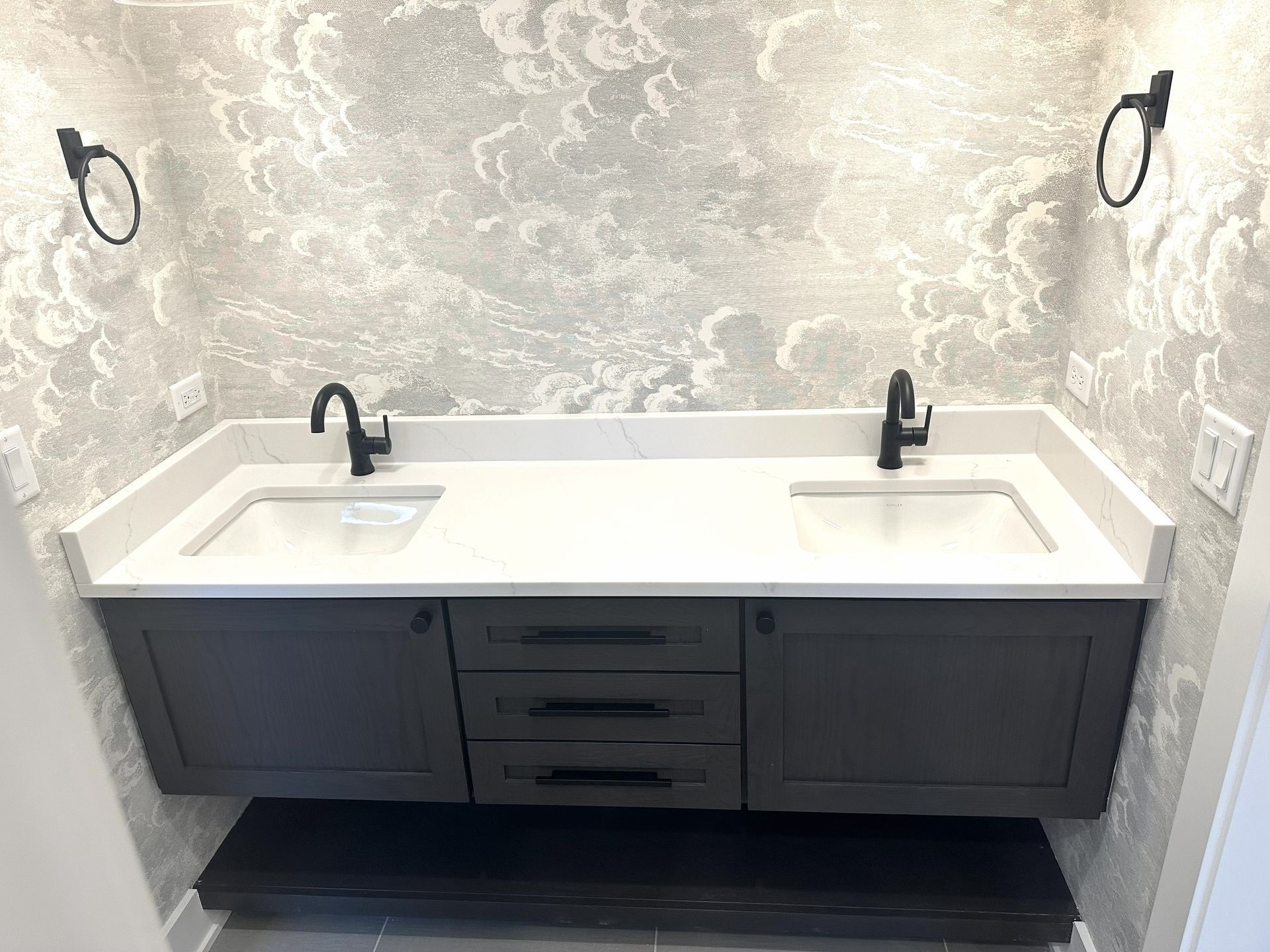 Modern bathroom vanity with double sinks, white countertop, and dark cabinets beneath patterned wallpaper.